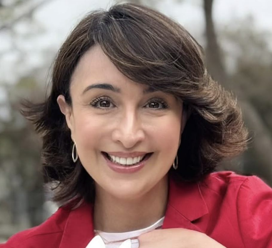 A woman with short brown hair and hoop earrings wearing a red blazer and white top, smiling outdoors.