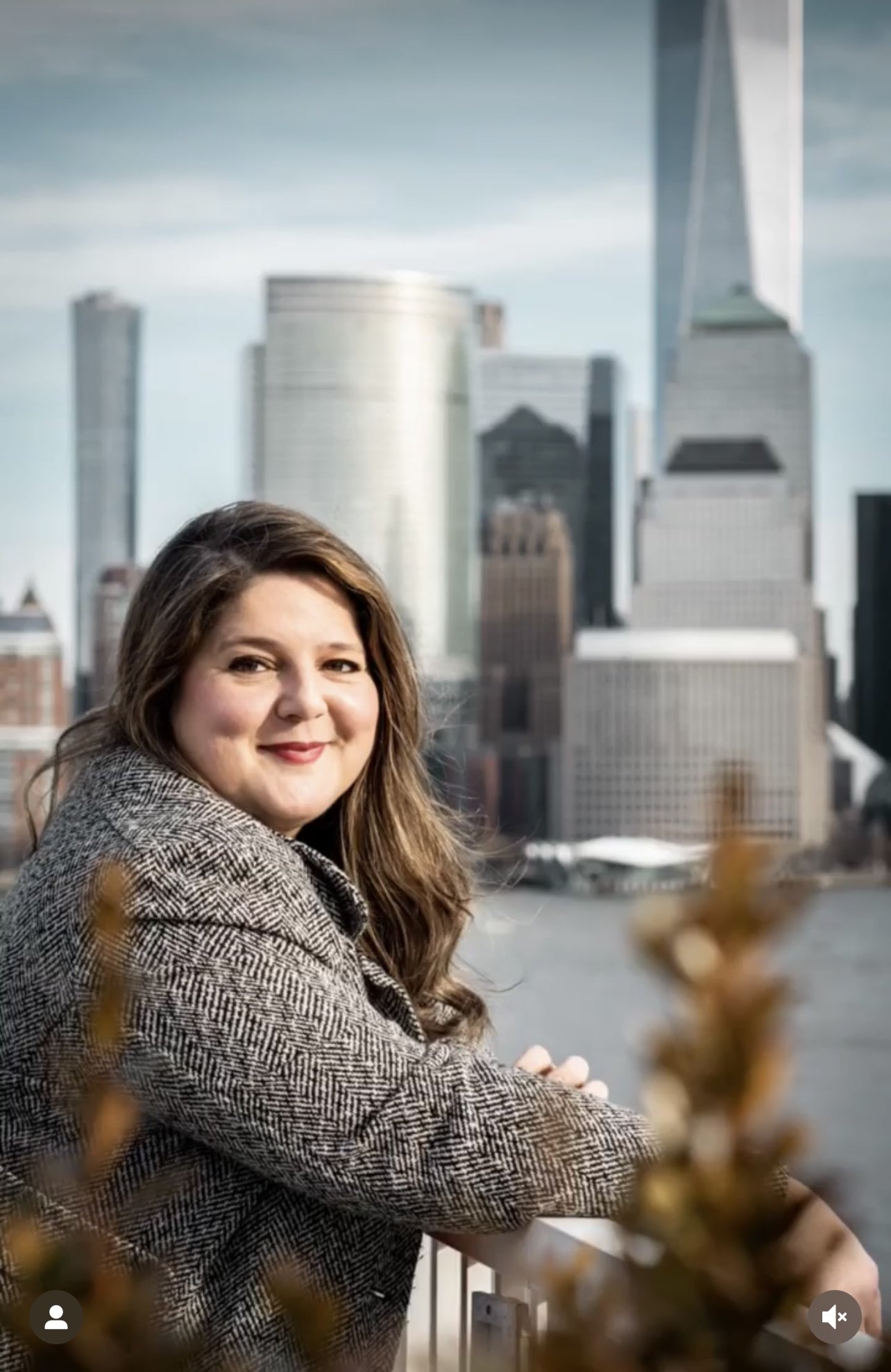 A woman with long brown hair sitting outdoors near a body of water in front of a city skyline with tall skyscrapers.