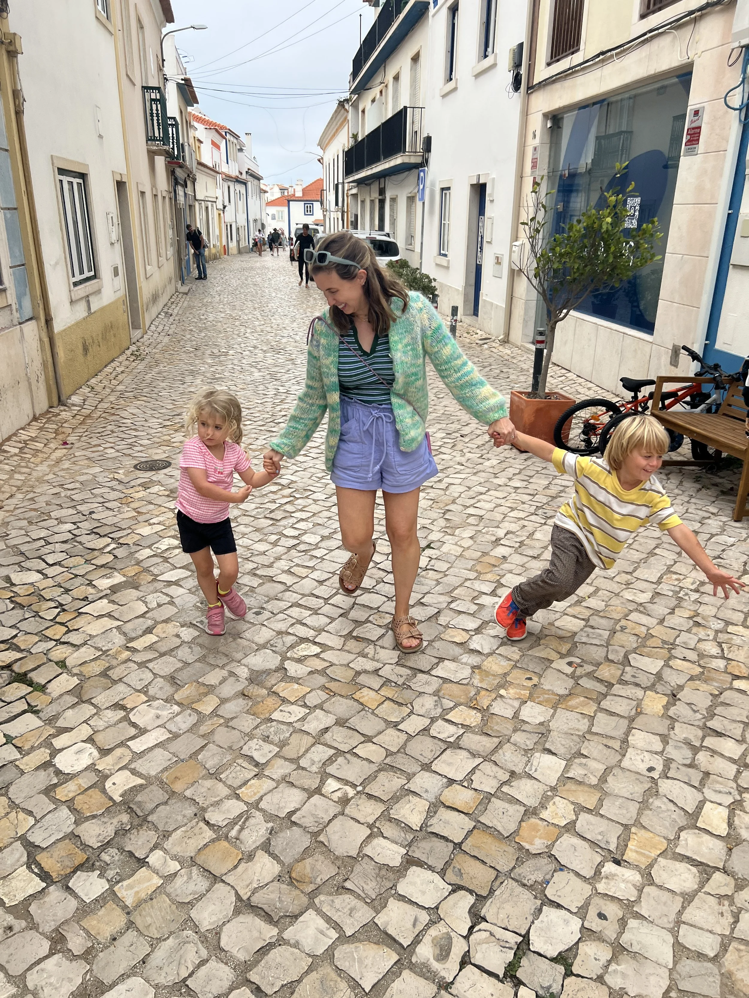 A woman holding hands with two children, one on each side, walking on a cobblestone street in a European town.