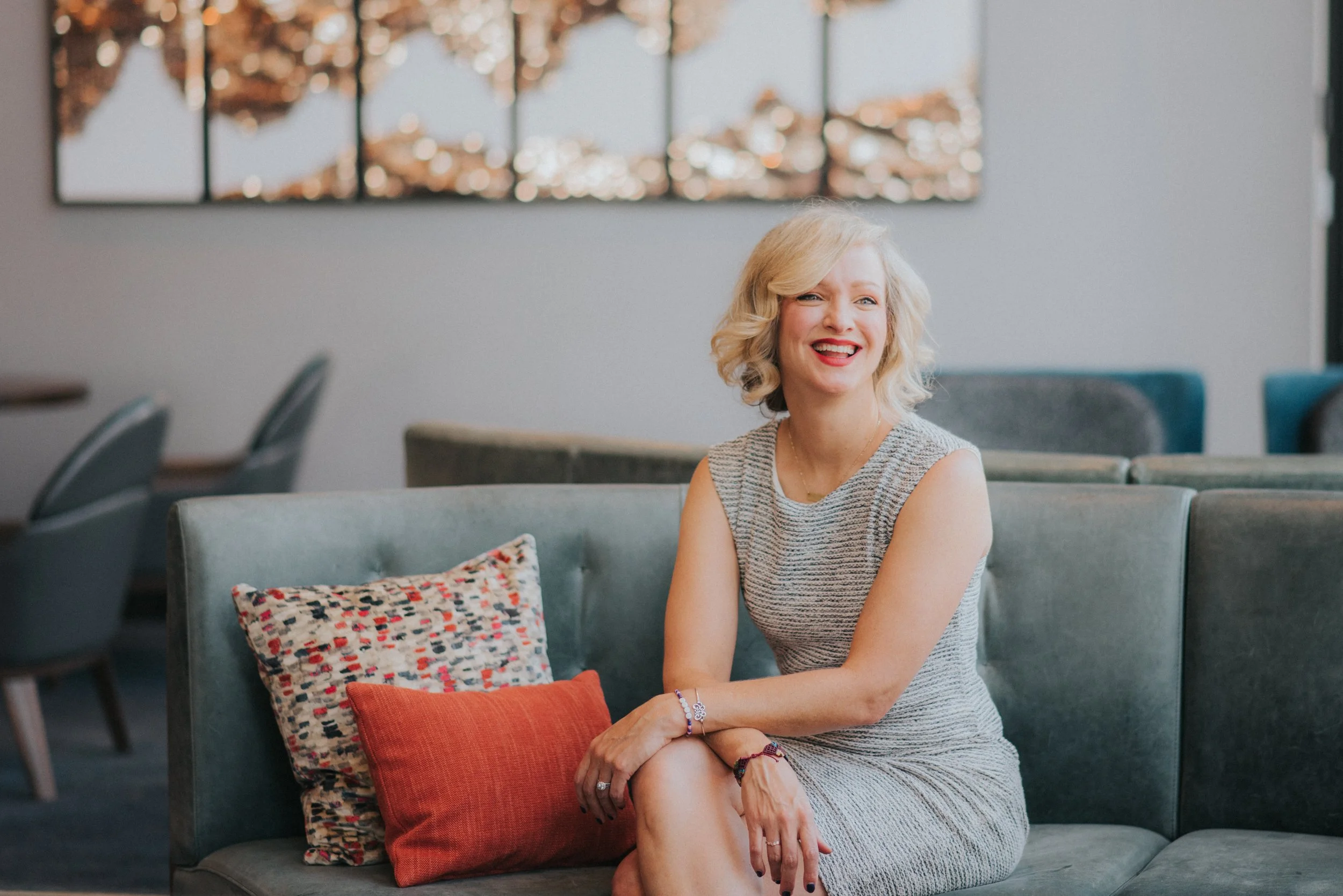 Vanessa Jupe, a woman with curly blonde hair, sitting on a gray sofa, smiling and laughing, in a modern indoor space with abstract wall art and chairs in the background.