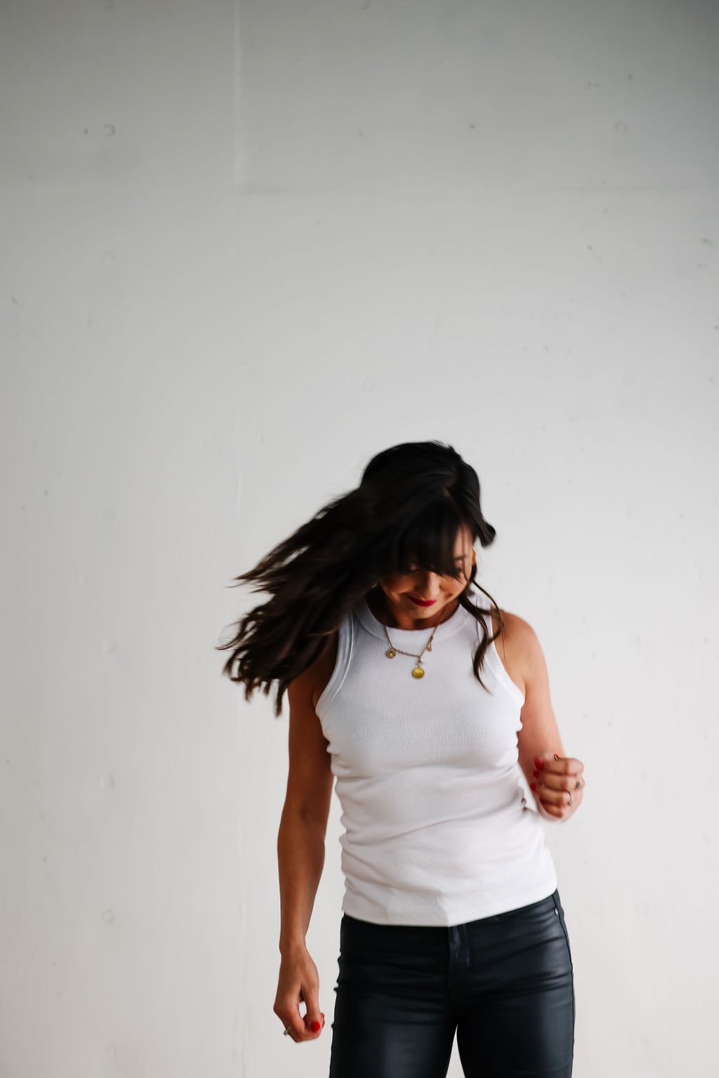 Woman with long dark hair dancing or moving, wearing a white sleeveless top and black pants, against a plain white wall.