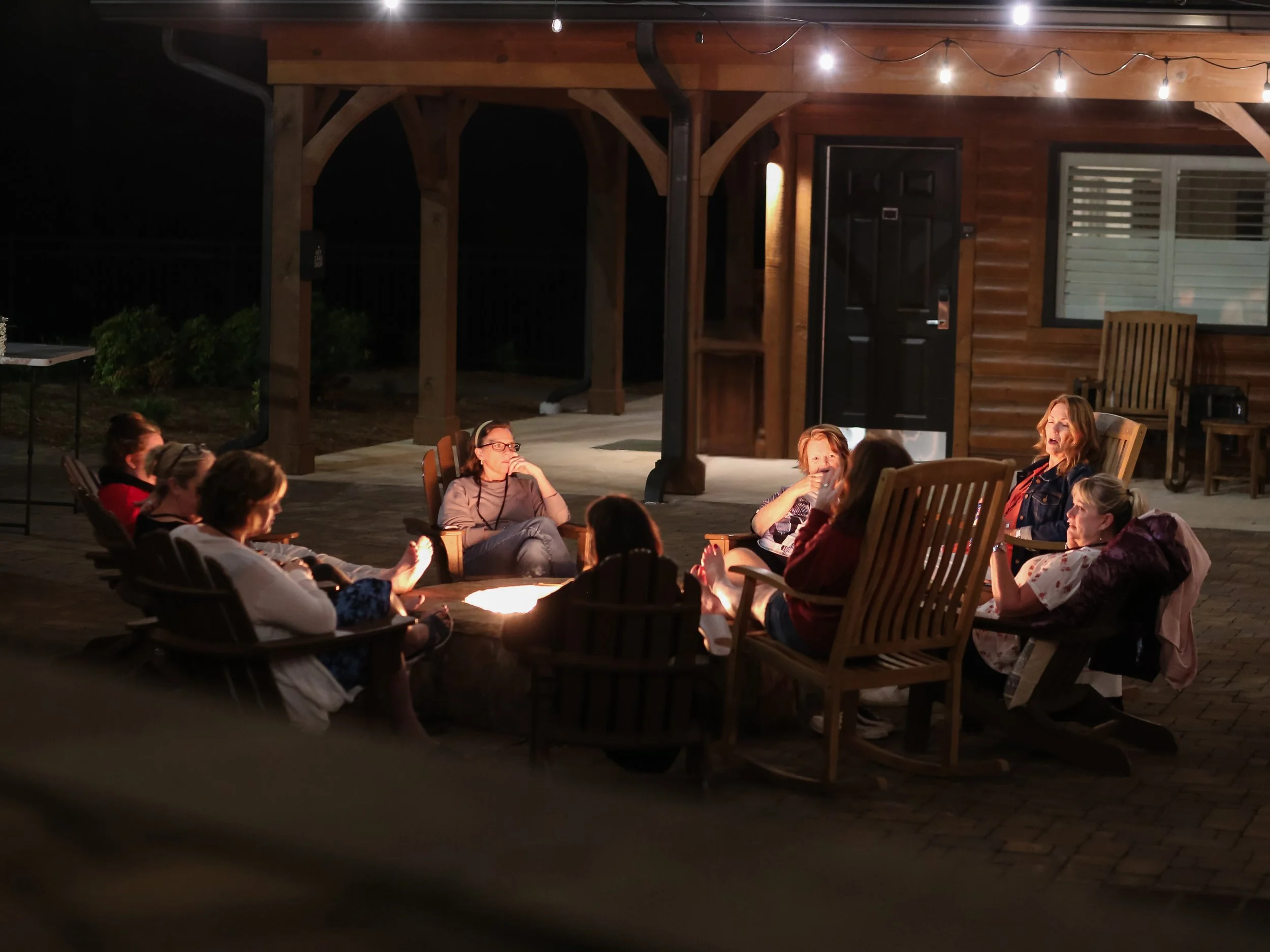 A group of retreat attendees sit in wooden chairs around a glowing fire pit, sharing a cozy evening under string lights on a porch.