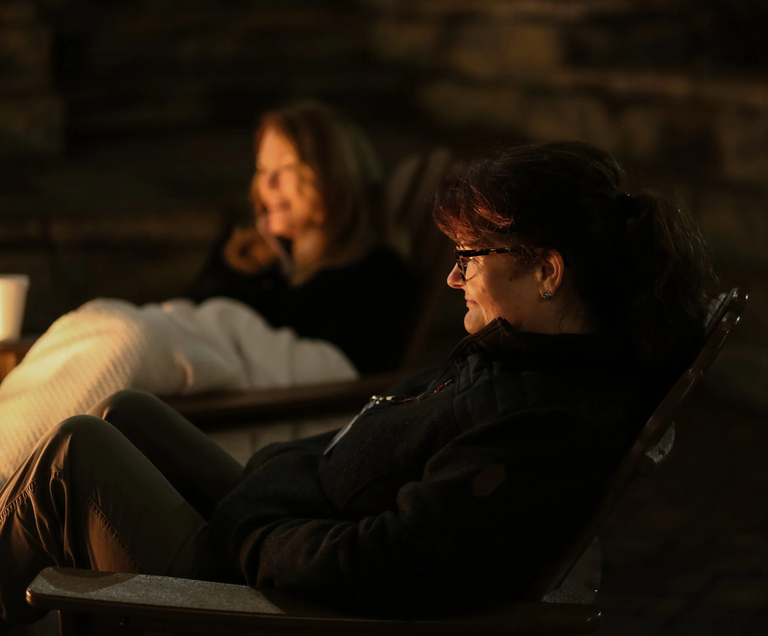 Two women sitting by the fire pit in the dark of night, the flames lighting up their faces