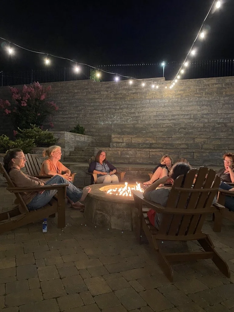 Several women sit and talk in Adirondack chairs around the fire pit at night under bistro lights