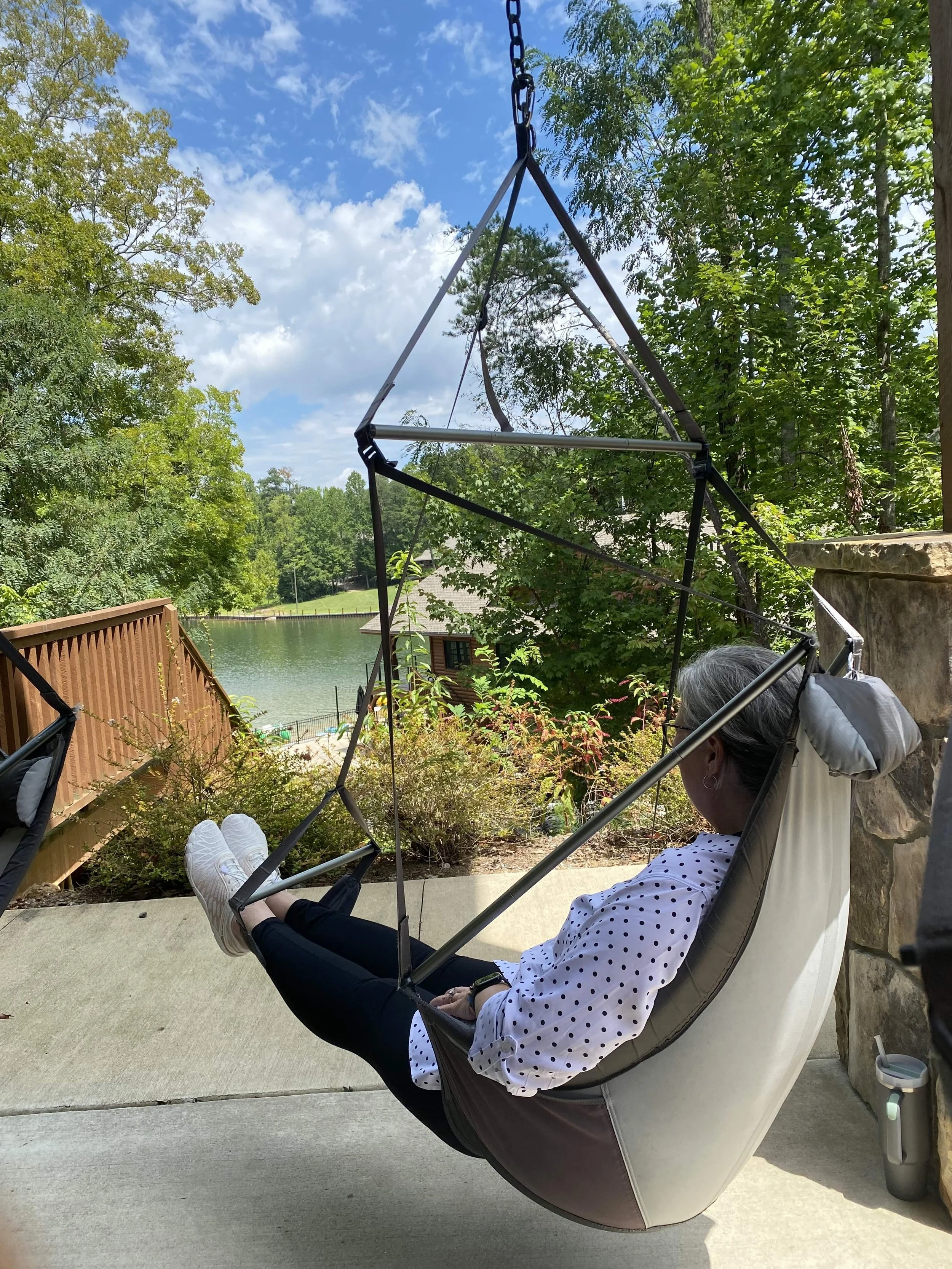 A retreat attendee relaxes in a hammock chair on a patio, overlooking a scenic lake surrounded by lush trees under a partly cloudy sky.