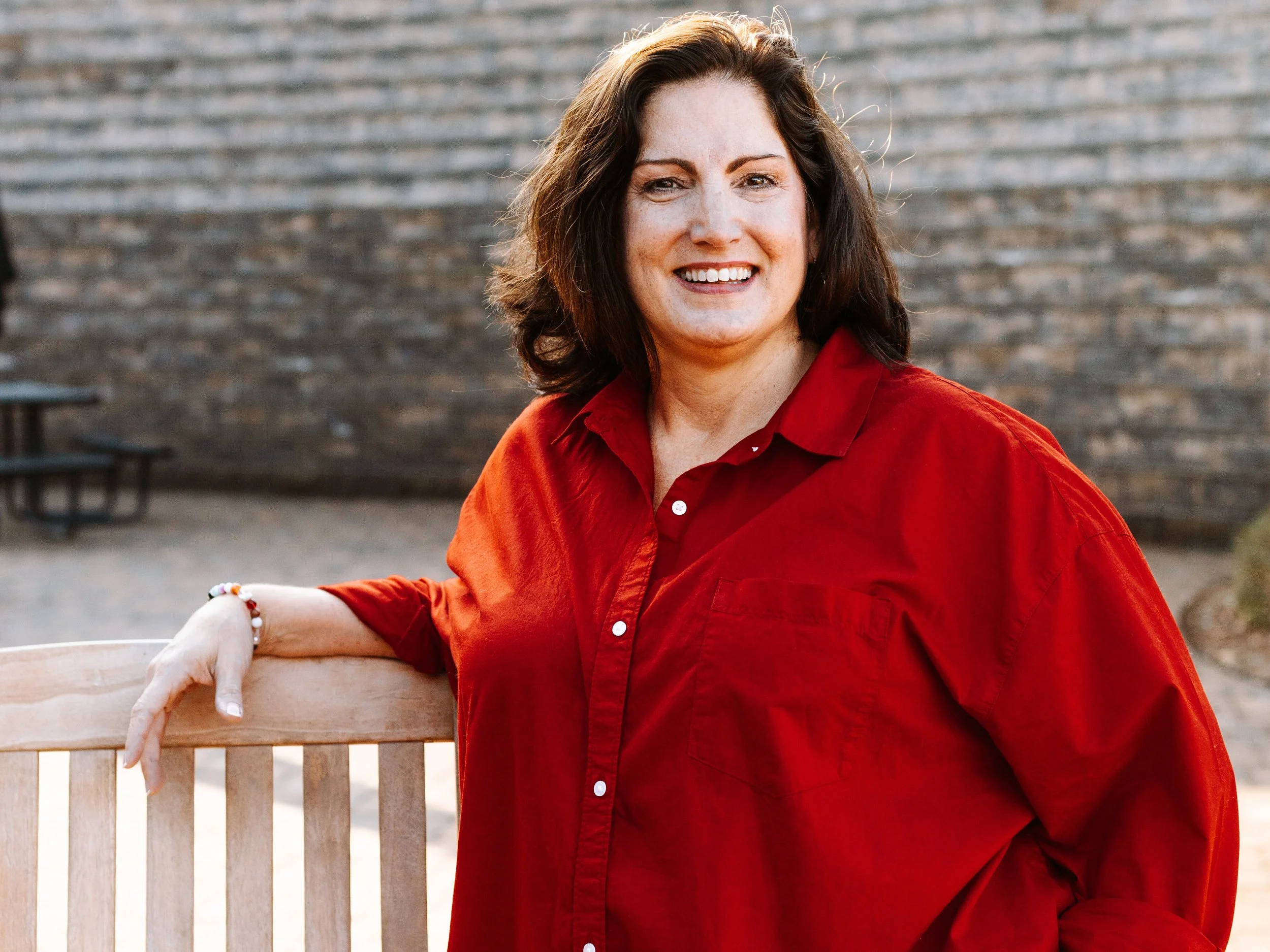 Nancy Squires smiles at the camera while wearing a red shirt and leaning on a wooden bench
