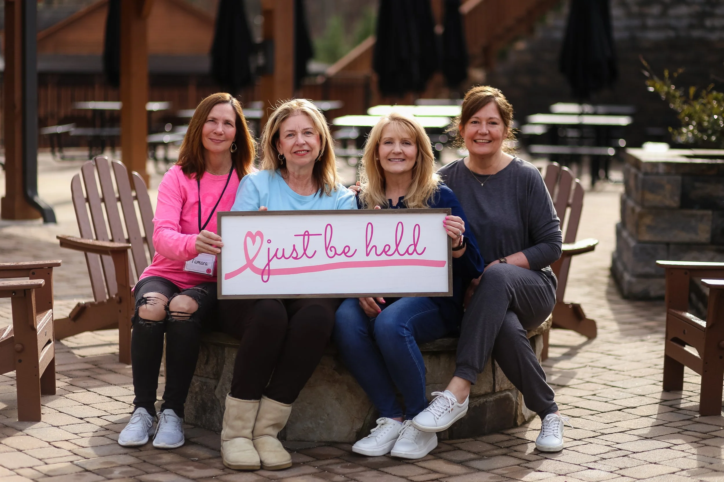 Four women sitting outside at retreat, holding a Just Be Held sign