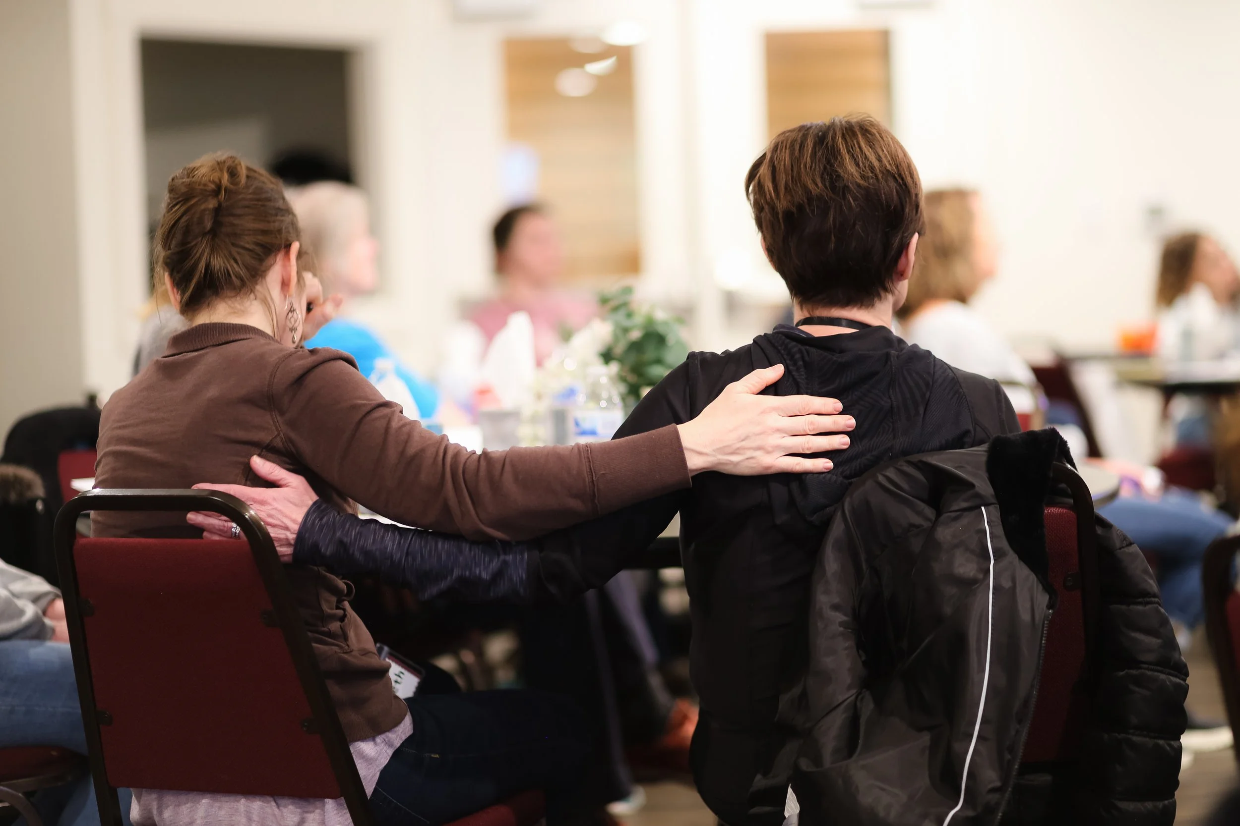 Two women sitting in the audience at retreat, and one has her hand on the other woman's back in a comforting way
