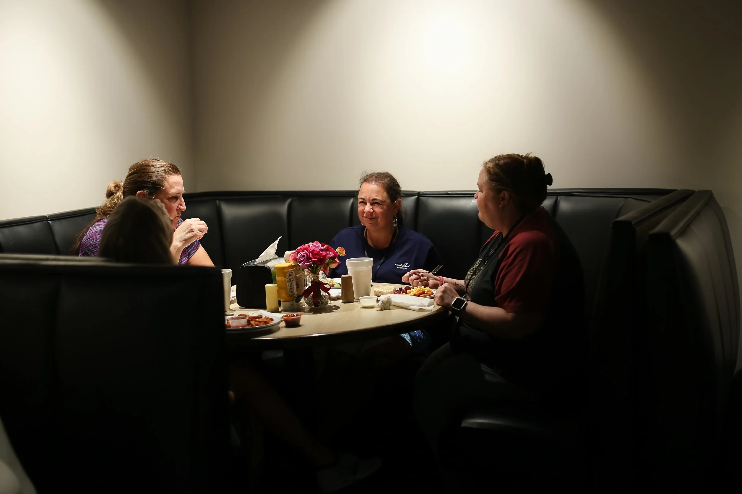 Four Just Be Held retreat attendees sit around a dimly lit booth in the dining room at Woodlands Camp, sharing food and conversation.