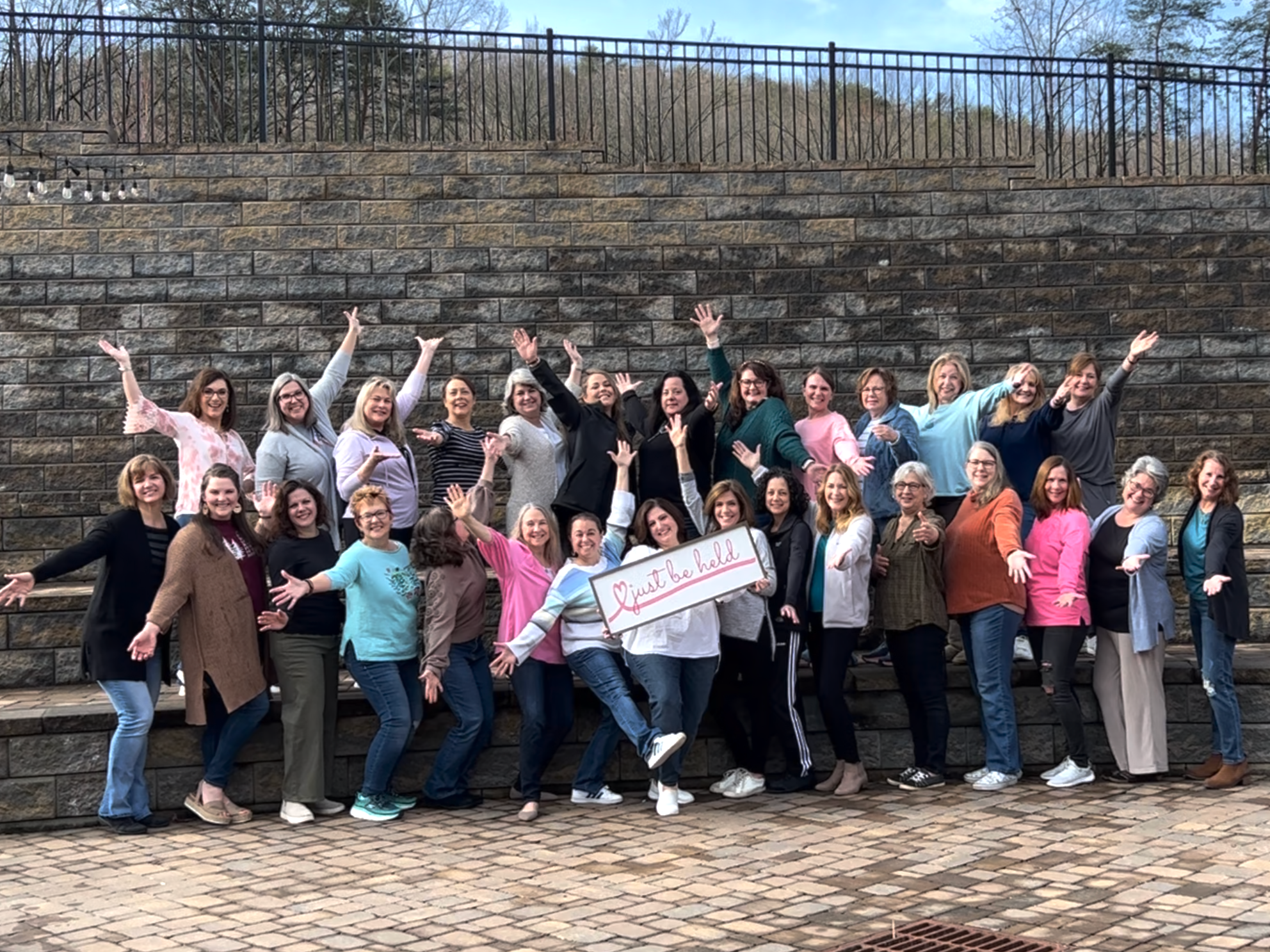 Around 30 women stand with arms open wide for a group photo, with Nancy Squires holding the Just Be Held sign in the middle of the group.