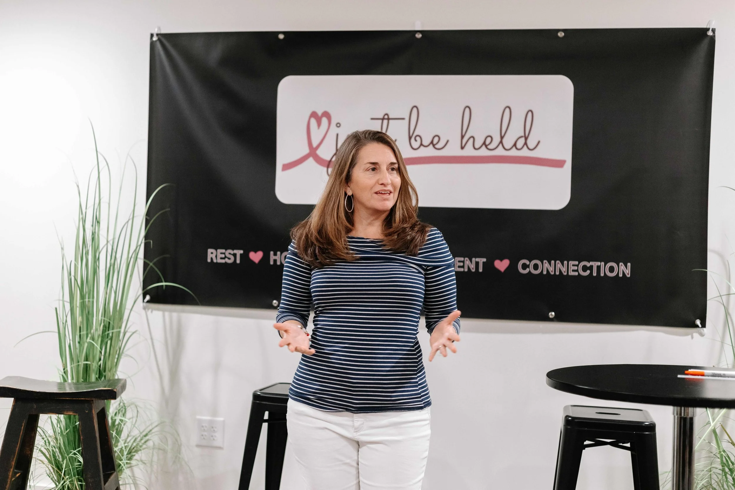 A woman stands in front of the Just Be Held sign and speaks to the women in attendance during Large Group time
