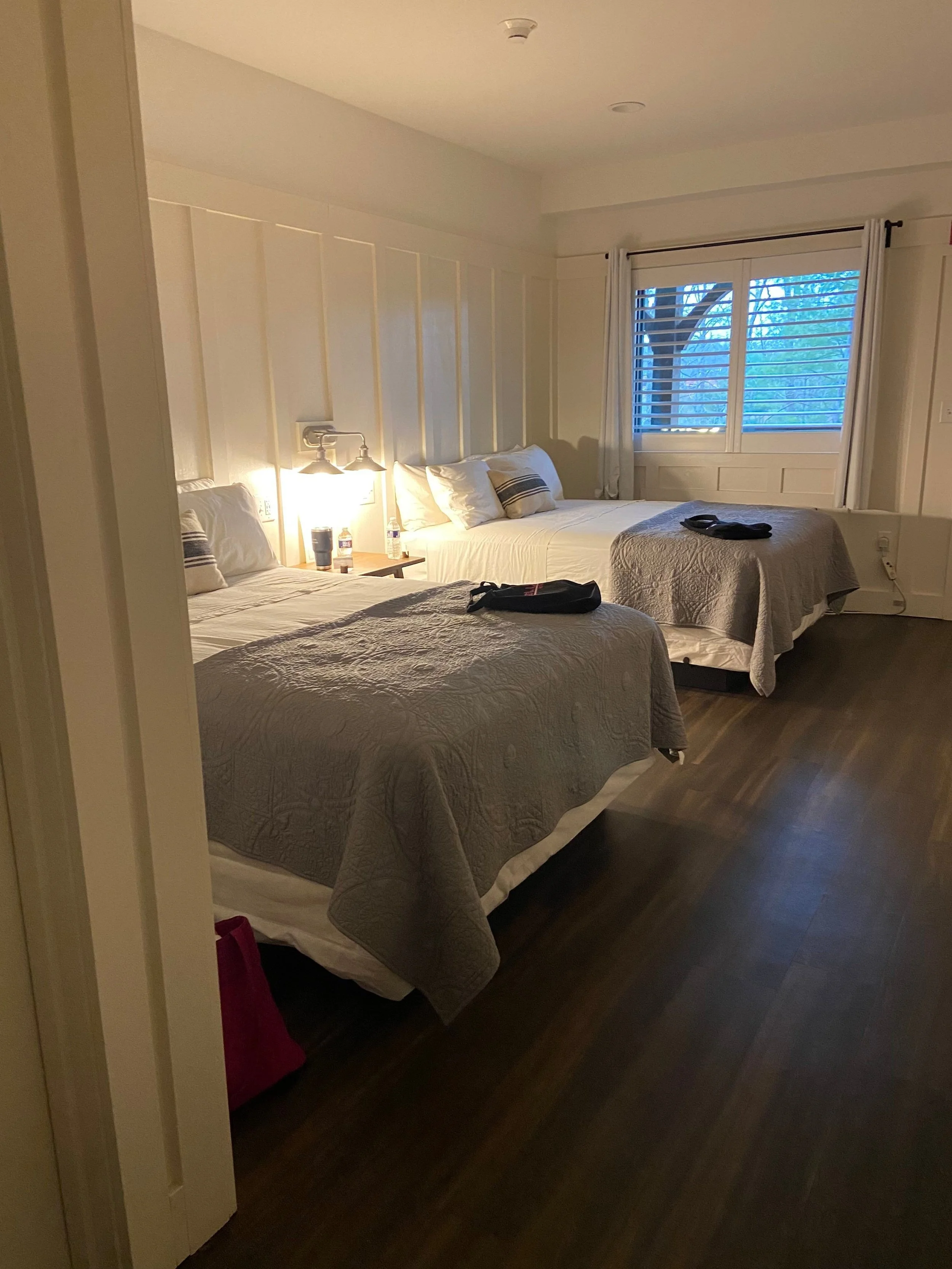 A cozy bedroom with two neatly made double beds, soft gray bedding, and pillows. Warm lighting from a wall lamp creates a relaxing ambiance.