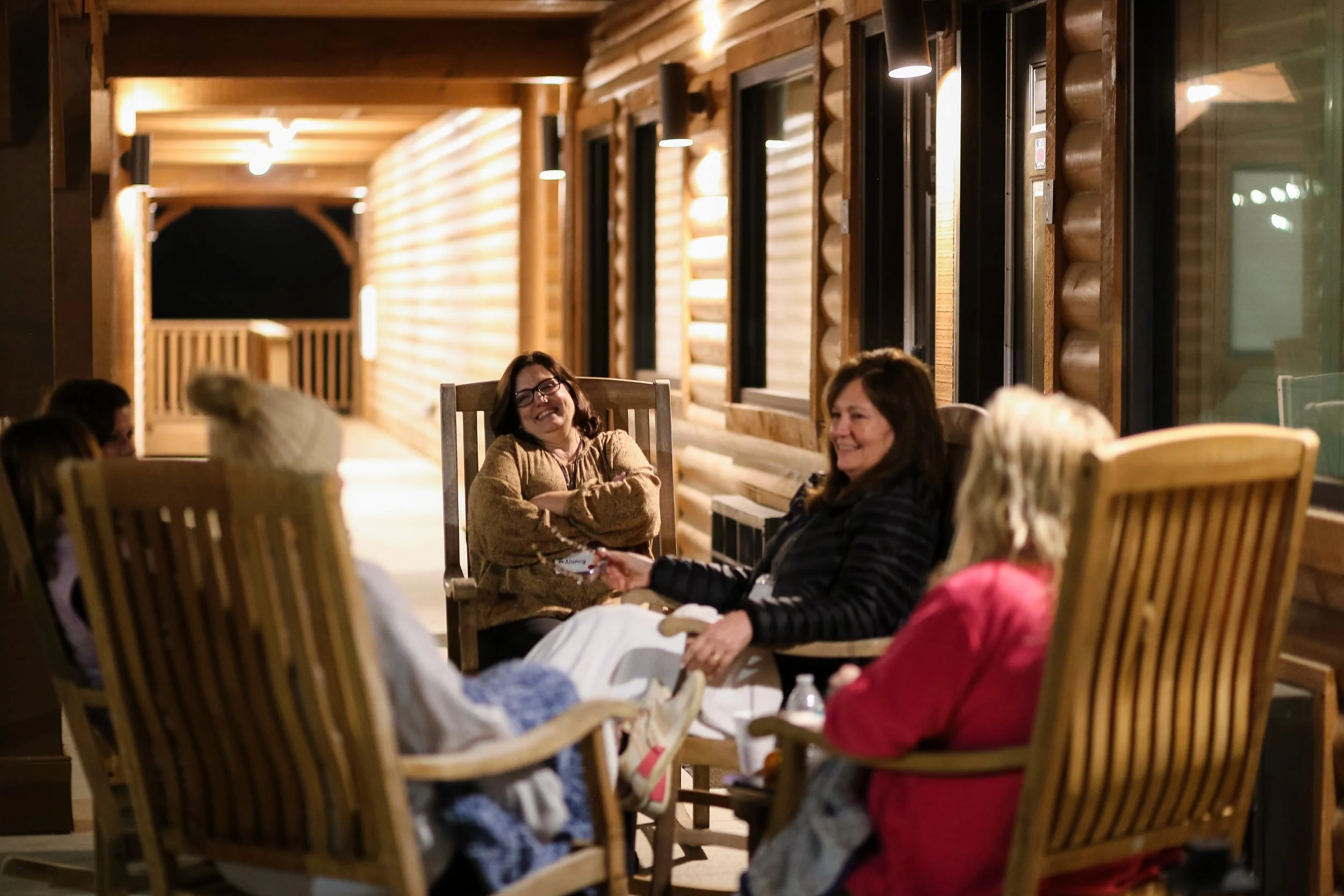 Rereat attendes sitting on wooden chairs on the porch of the Lodge at night, smiling and chatting.
