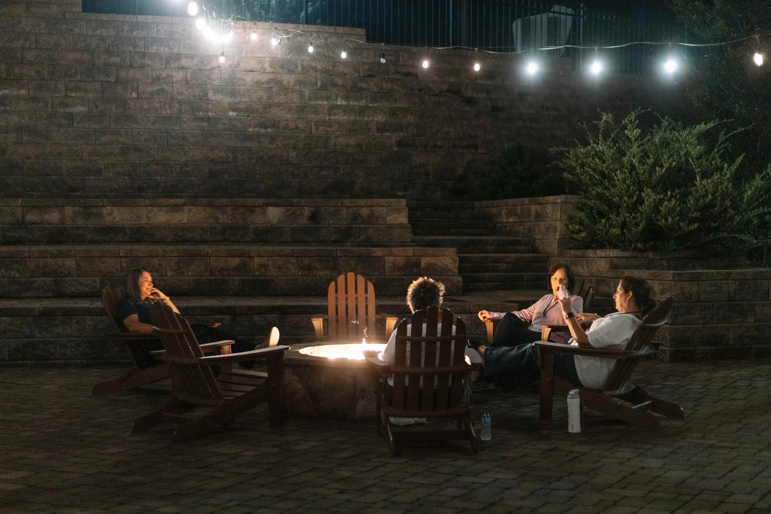 Four women sitting in Adirondack chairs around the fire pit at night, with bistro lights hanging above