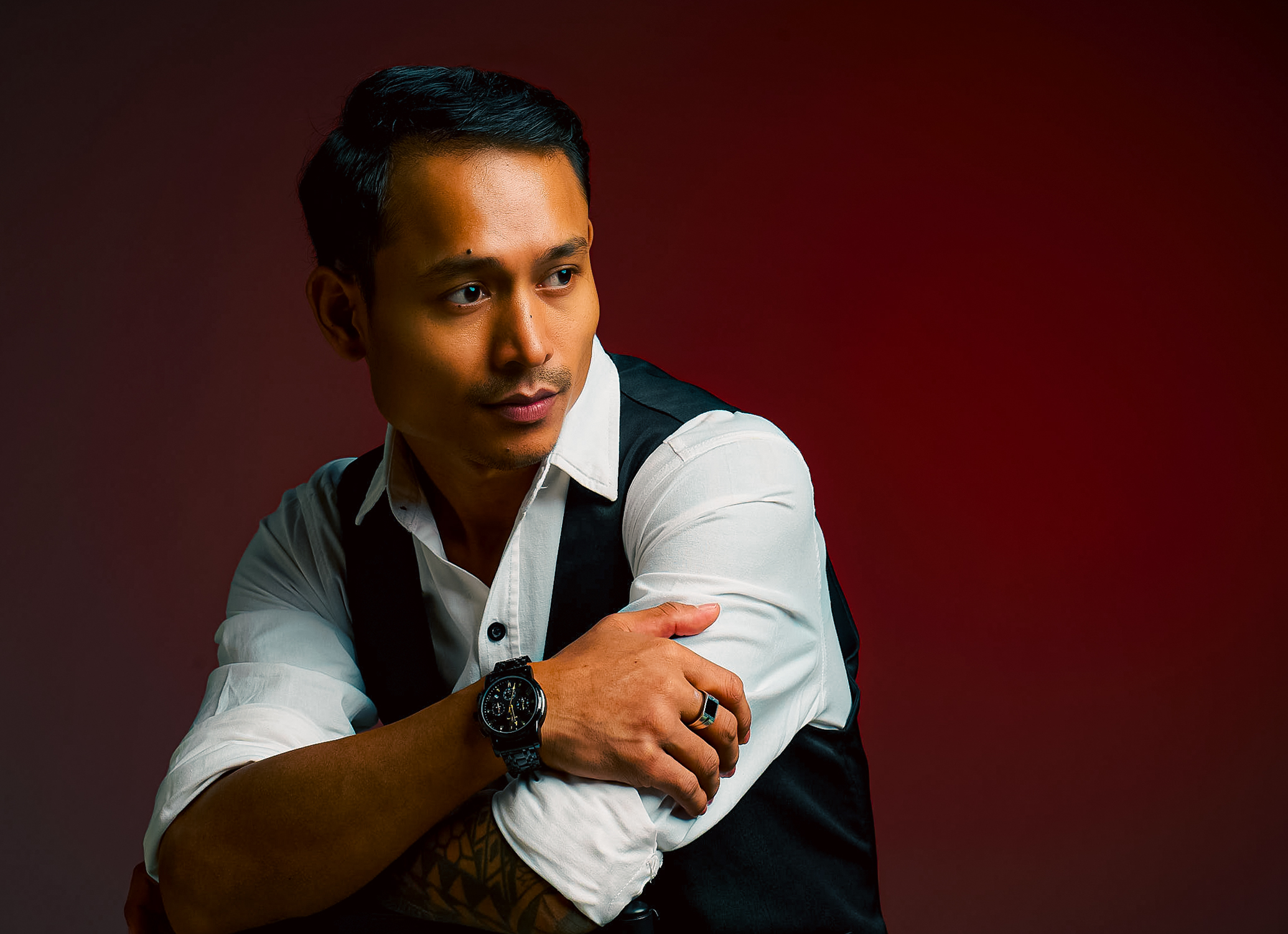 Portrait of a young man with a dark, styled haircut, wearing a white dress shirt with sleeves rolled up, a black vest, glasses, a wristwatch, and a ring, posing against a red gradient background.