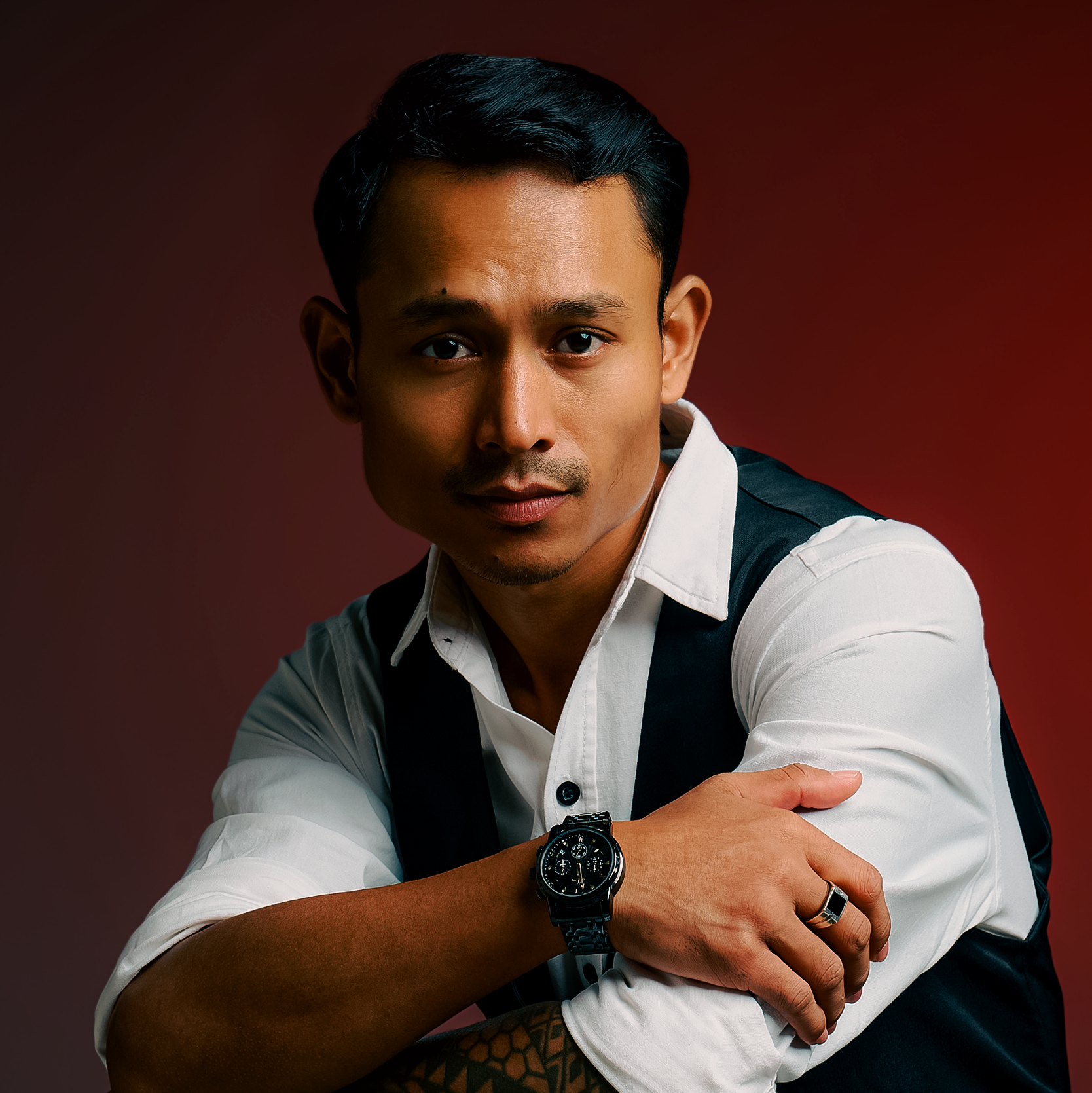 A young man with black hair, wearing a white shirt with rolled sleeves, a black vest, and a large wristwatch on his left wrist, looking directly at the camera with a serious expression.
