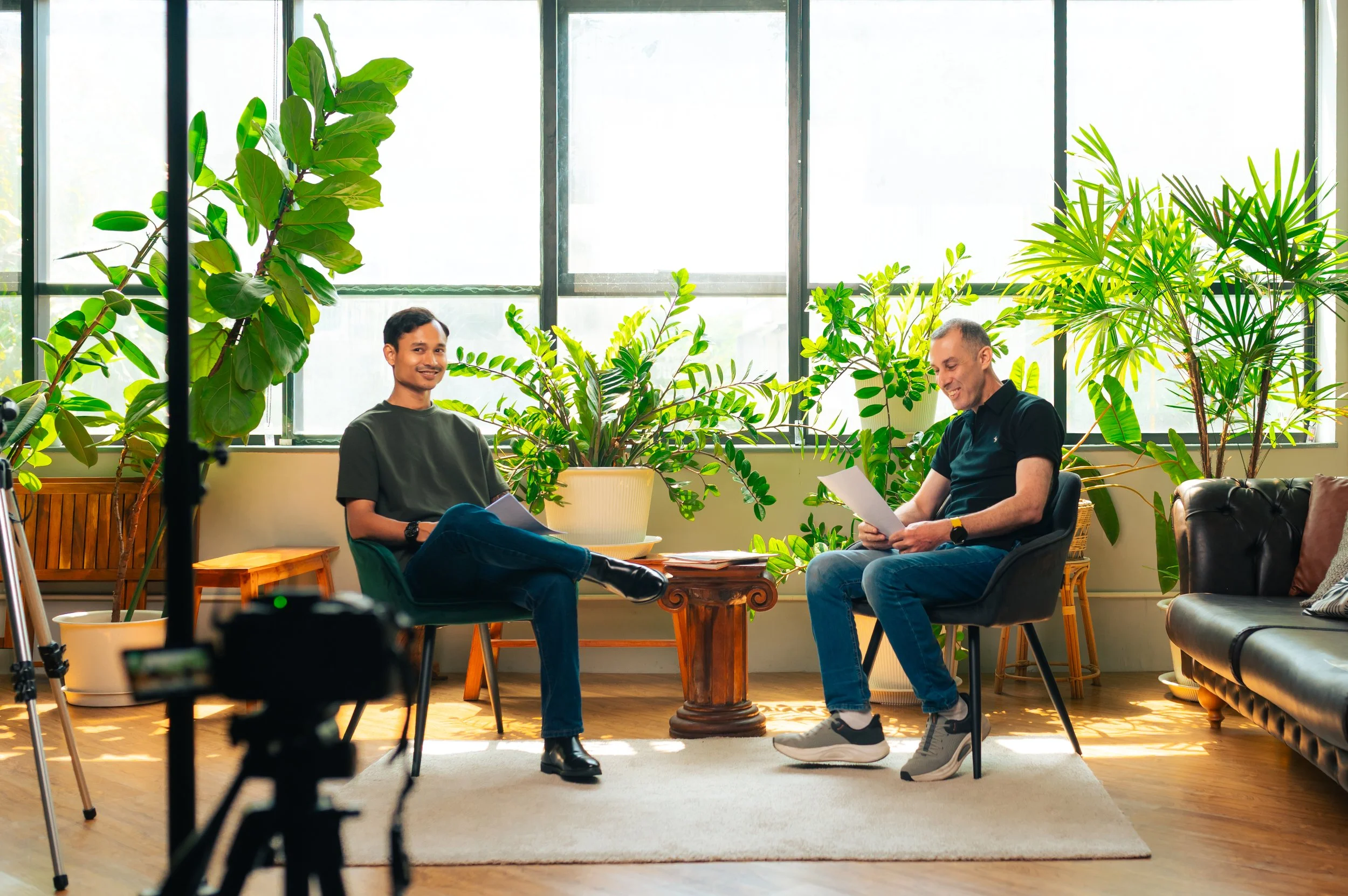 Two men sitting in a bright room with large windows and green plants, engaged in a conversation with papers, with a camera in the foreground.