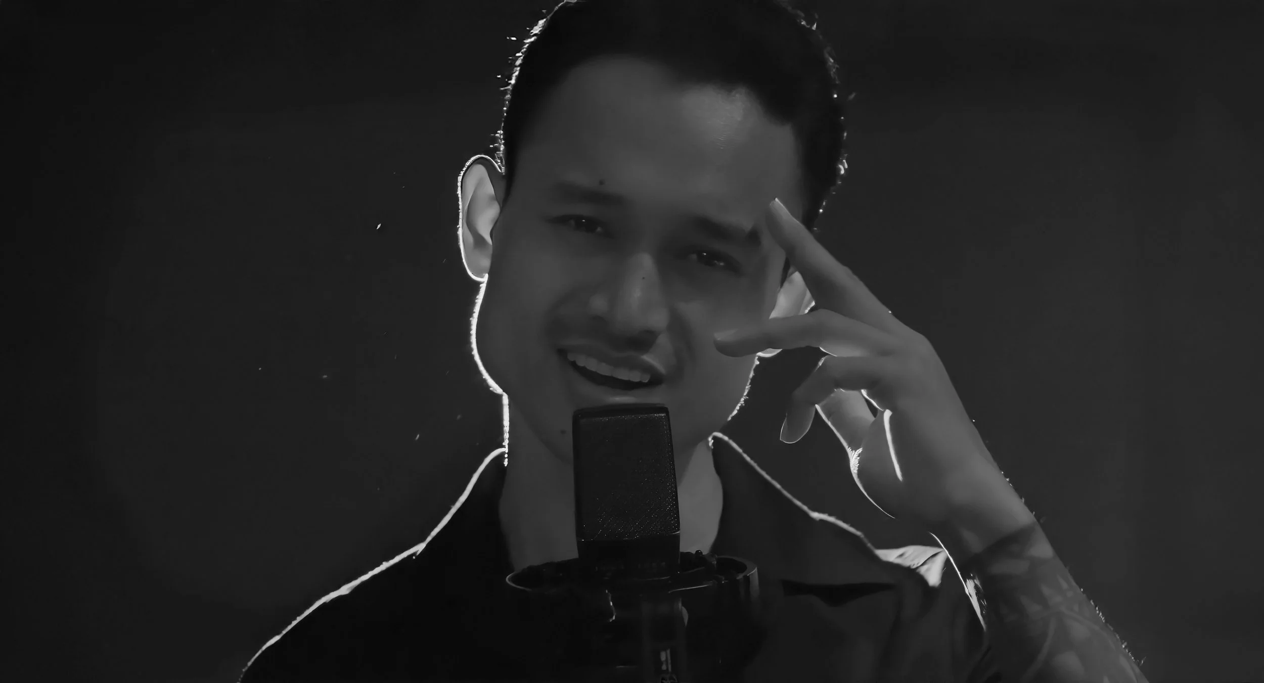 A young man in a black shirt singing into a microphone, touching his temple with his right hand, illuminated by backlight, in black and white.