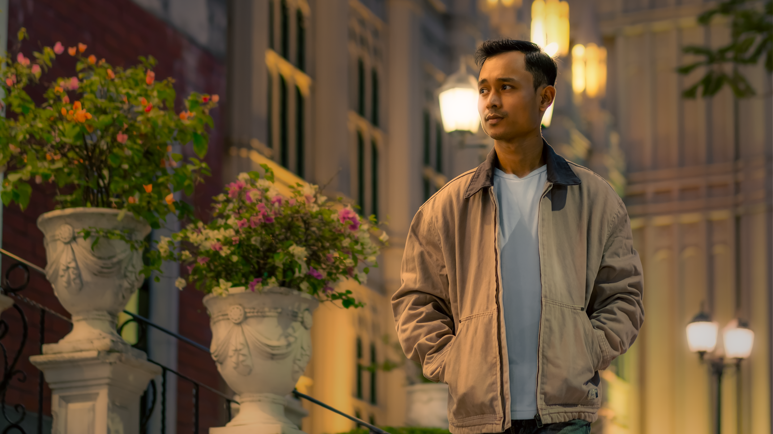 A man walking on a city street at dusk, wearing a beige jacket and white shirt, with decorative flower pots on a railing nearby.