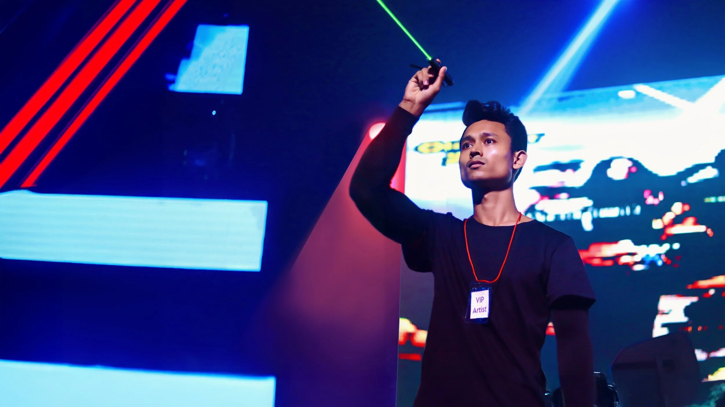 Young male artist on stage, wearing a VIP badge, holding a green laser pointer, with colorful digital screens in the background.