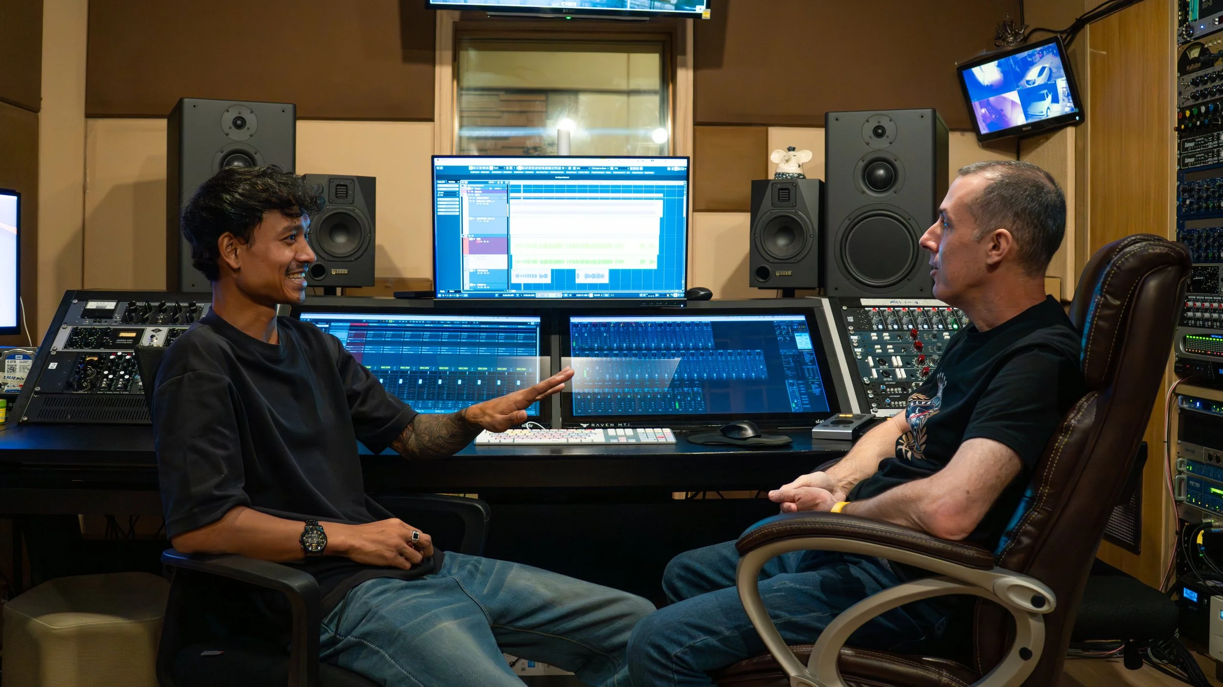 Two men conversing in a recording studio with large speakers and multiple computer screens displaying audio editing software.