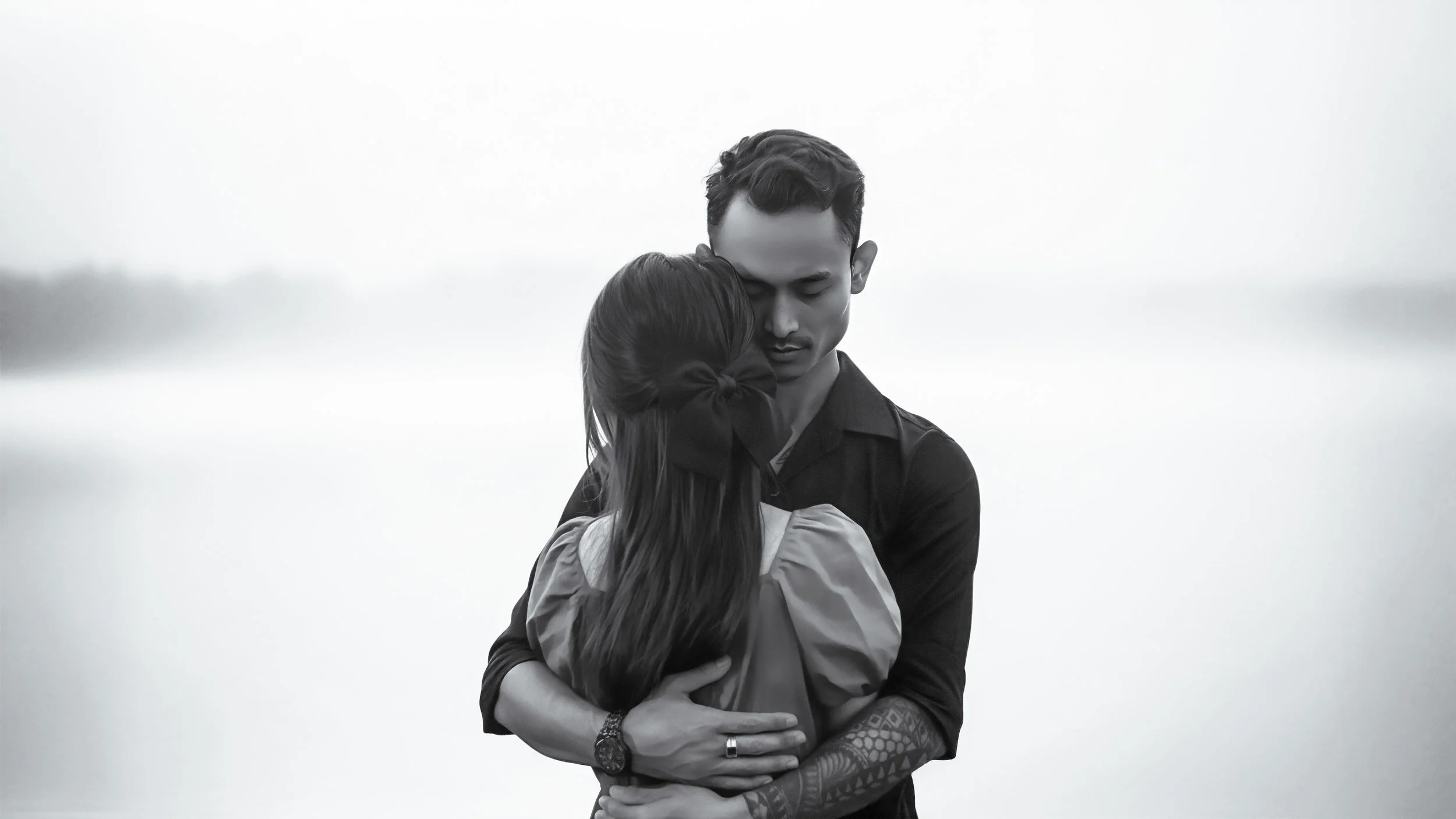 A black-and-white photo of a couple hugging each other closely outdoors. The man has short dark hair, tattoos on his arm, and is wearing a dark shirt. The woman has long dark hair tied back with a large bow, and is wearing a light-colored top. They are standing near a body of water, with a calm and serene background.