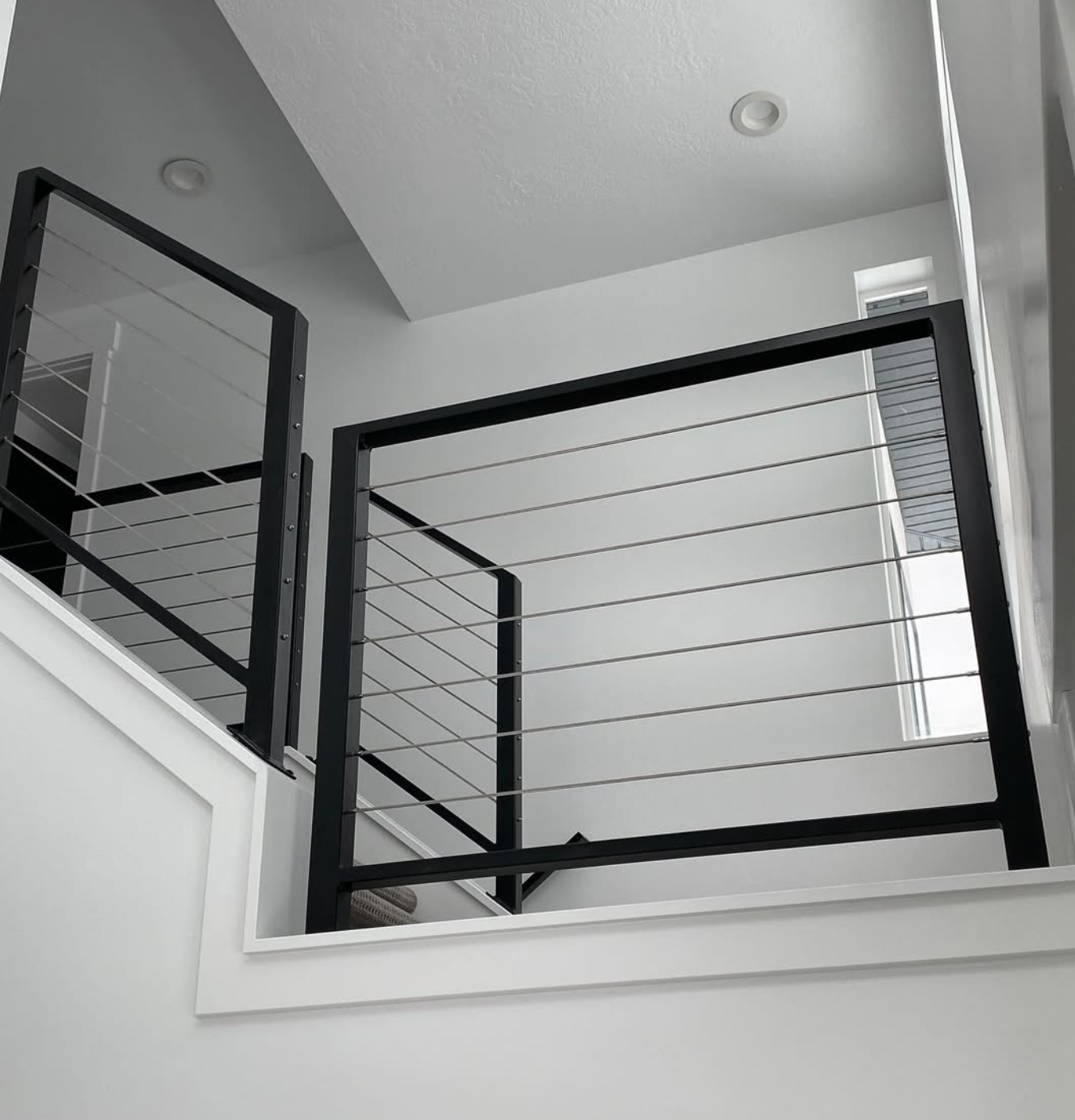 Interior view of a modern, minimalistic staircase with black metal railings and horizontal metal cables, white walls, ceiling lights, and a small window