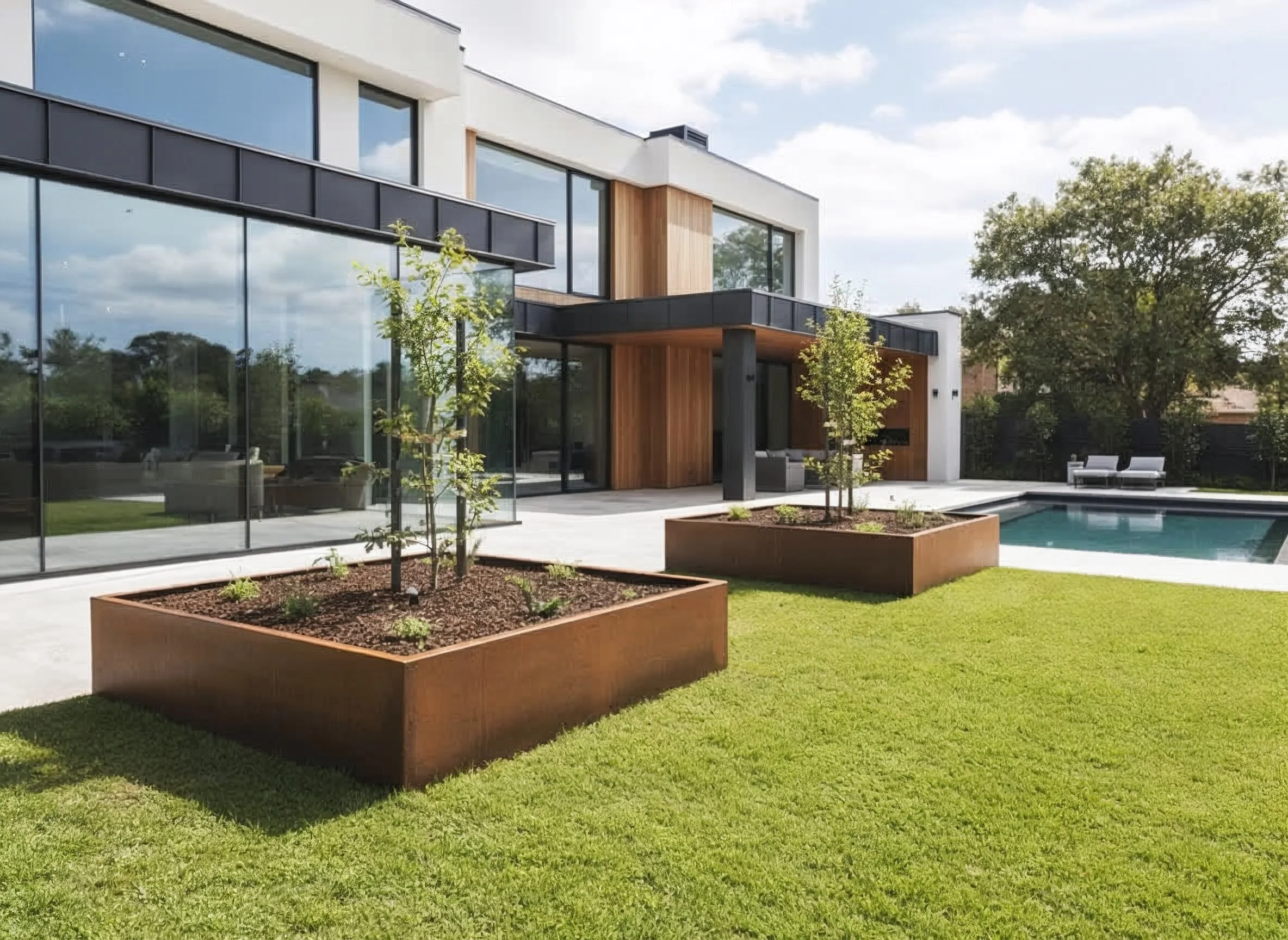 Modern house with large glass windows, a patio, two planters with small trees, a swimming pool, and outdoor seating, surrounded by green grass and trees.