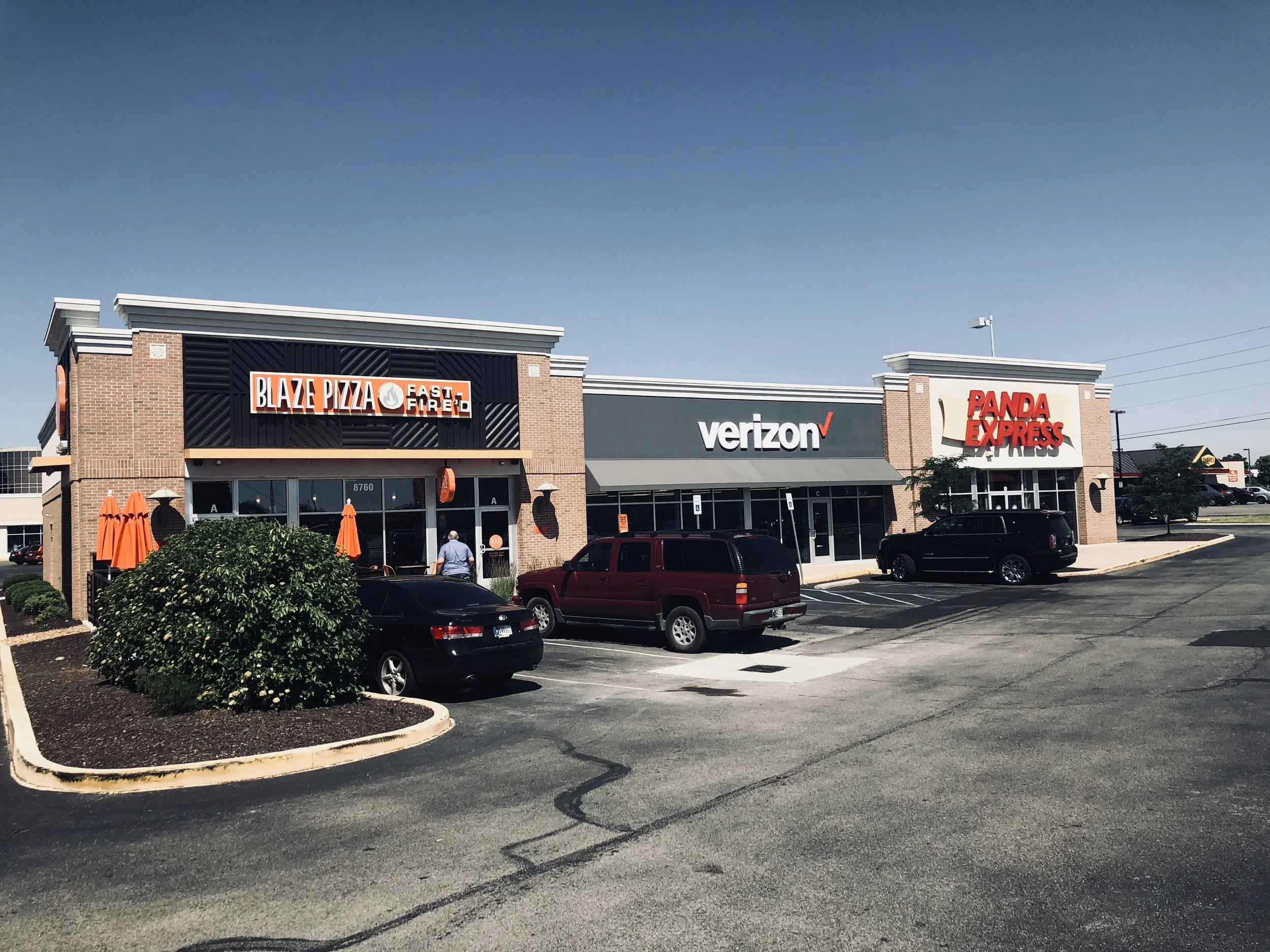 A shopping plaza parking lot with several parked cars and storefronts. The stores include Blaze Pizza, Verizon, and Panda Express. There are orange umbrellas and a small bush in the foreground. The sky is clear and blue.