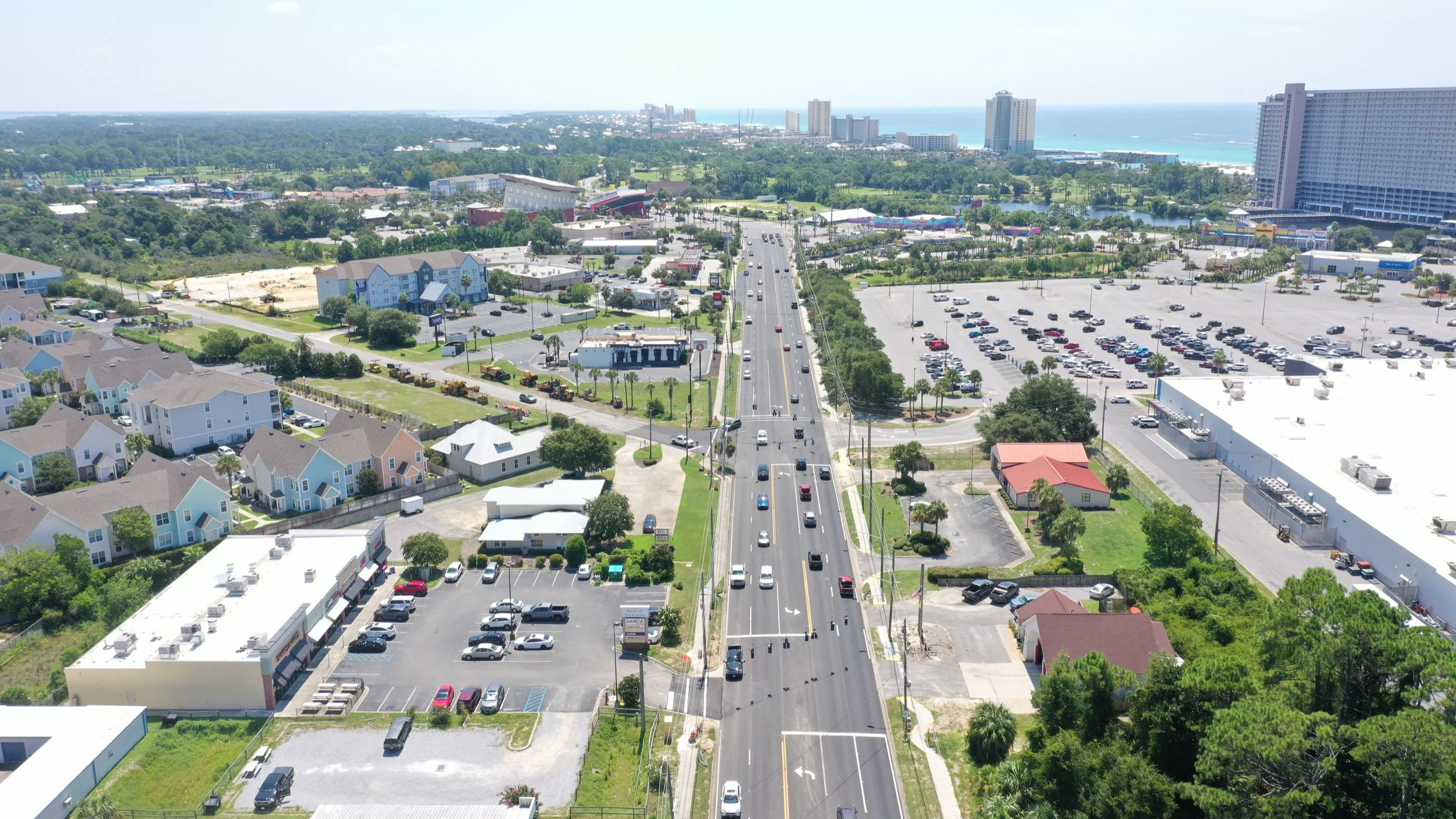 Aerial view of a city with a main road running through, lined with cars, surrounded by residential and commercial buildings, green trees, and a coastline with high-rise hotels and the ocean in the background.