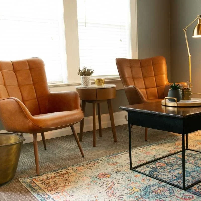 Living room with two tan leather armchairs, a small round side table with plants and a candle, a black coffee table with decorative items, a gold floor lamp, and a patterned area rug near windows with closed blinds.