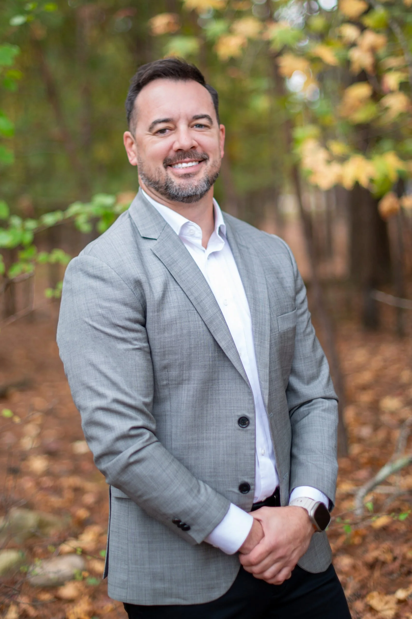 A man in a gray suit smiling outdoors in a wooded area with autumn leaves.