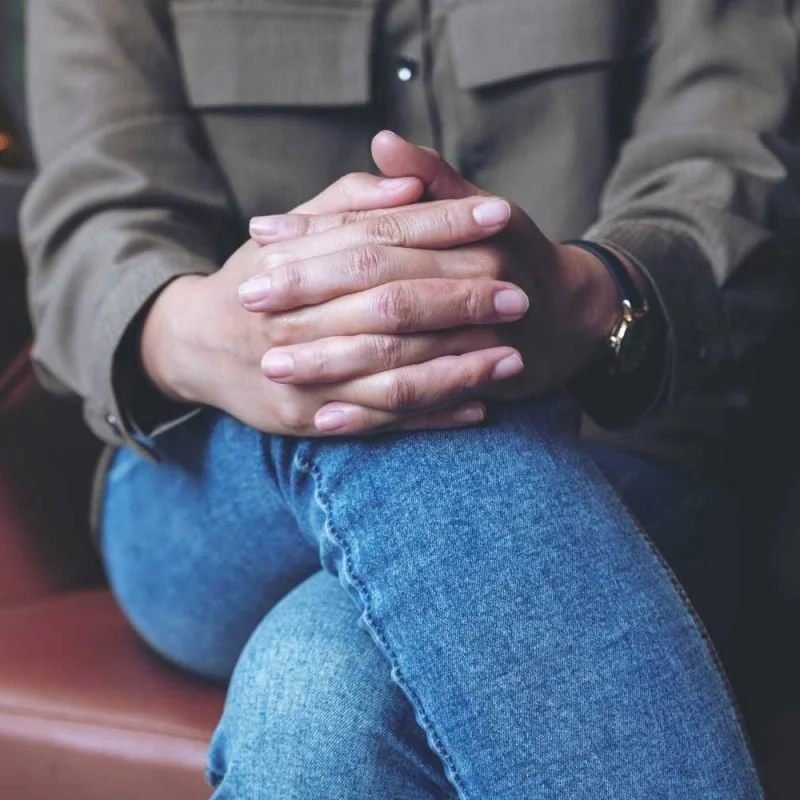 Person sitting with crossed legs, hands clasped together on top of the knee, wearing a brown jacket, blue jeans, and a wristwatch.