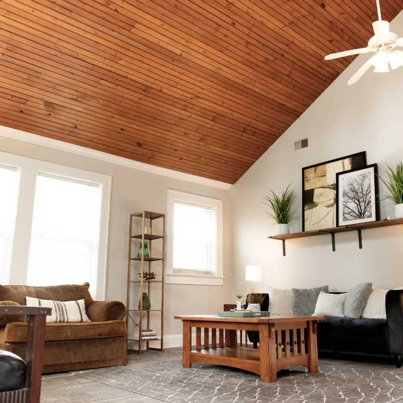 Living room with wooden vaulted ceiling, white walls, three windows, a brown sofa, a black sofa, a wooden coffee table, a tall bookshelf, framed pictures, houseplants, and a ceiling fan.