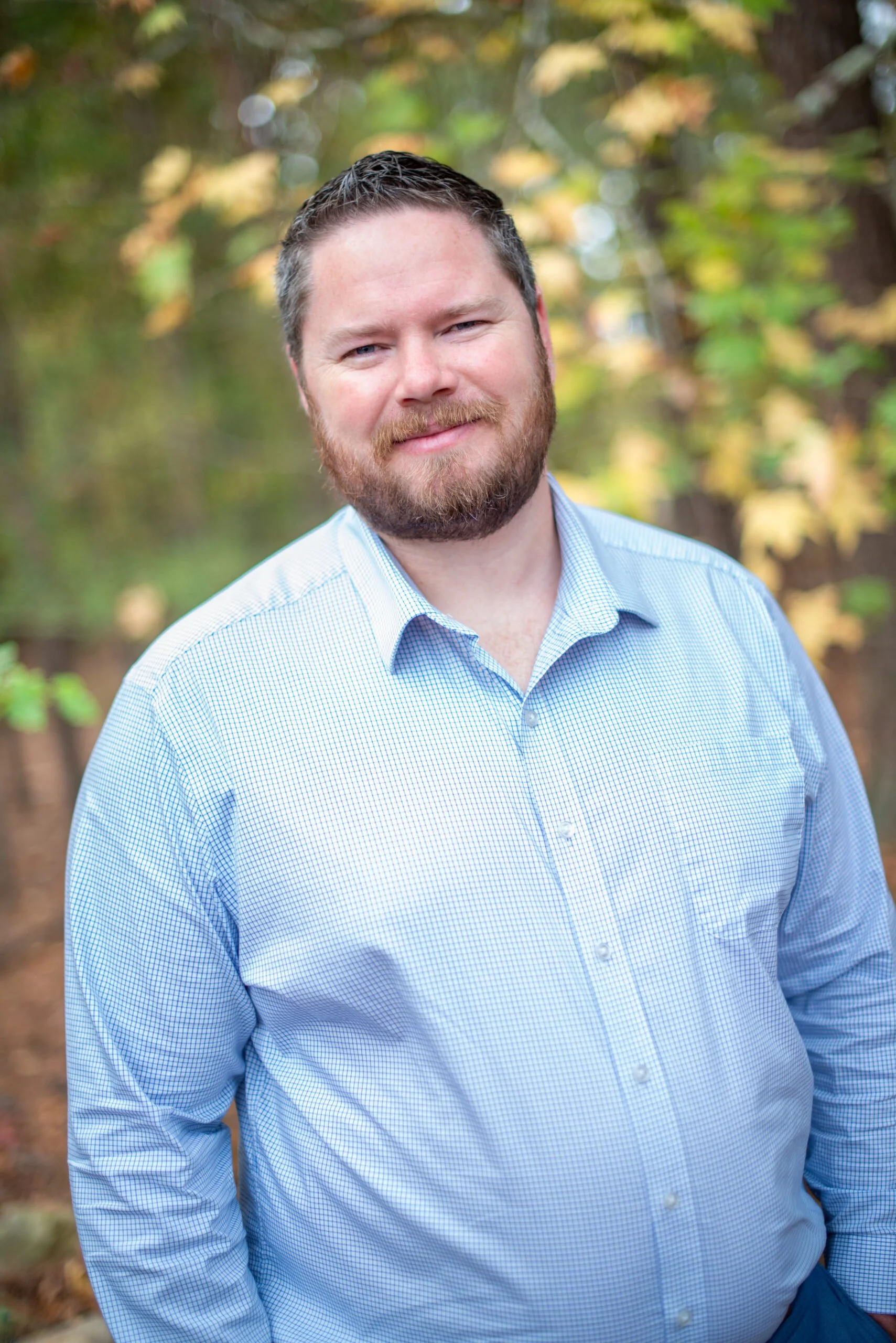 A man with a beard and short brown hair wearing a light blue checkered shirt, standing outdoors with blurred trees in the background.