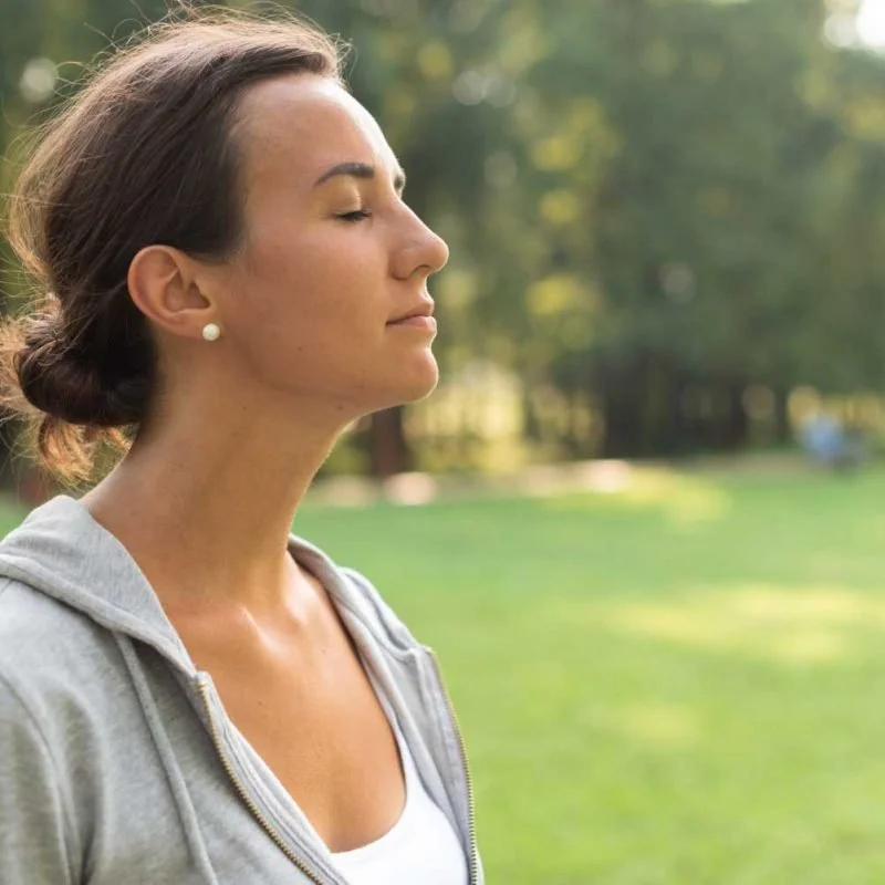 A woman outdoors with her eyes closed, practicing meditation or mindfulness, wearing a gray hoodie and white top, with a background of green grass and trees.