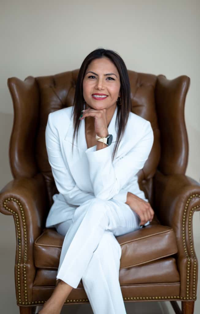 A woman dressed in a white suit sitting on a brown leather armchair, smiling with her chin resting on her hand.