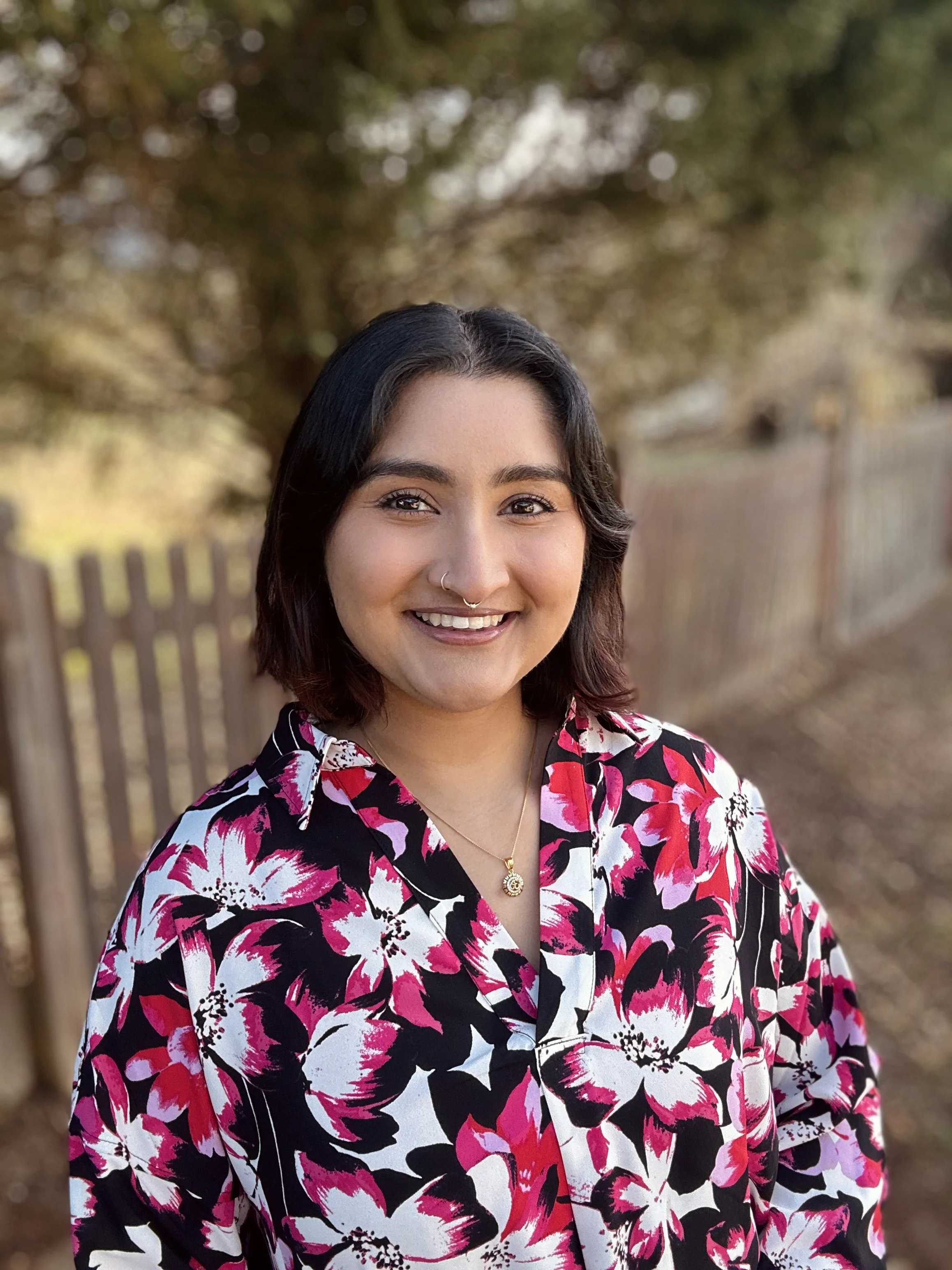 Portrait of a young woman outdoors with trees and a wooden fence in the background, wearing a black, white, and pink floral shirt, gold necklace, and nose ring, smiling at the camera.