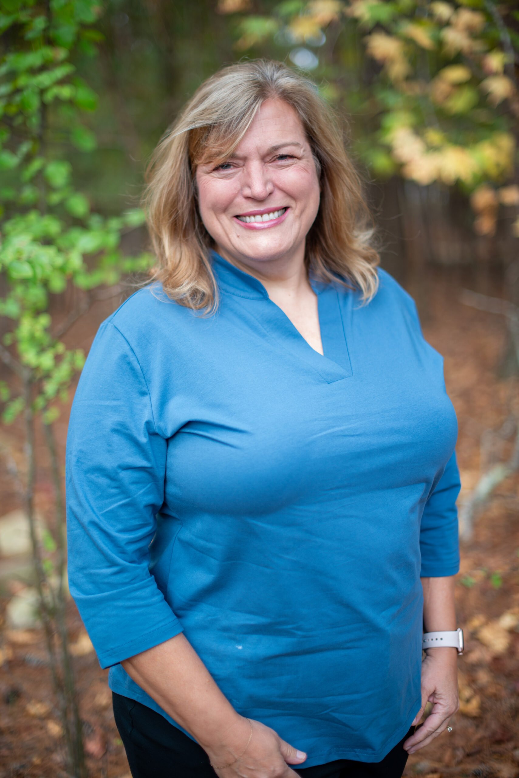 A smiling woman with blonde hair is standing outdoors in a park, wearing a blue top and a white watch, with greenery and autumn leaves in the background.