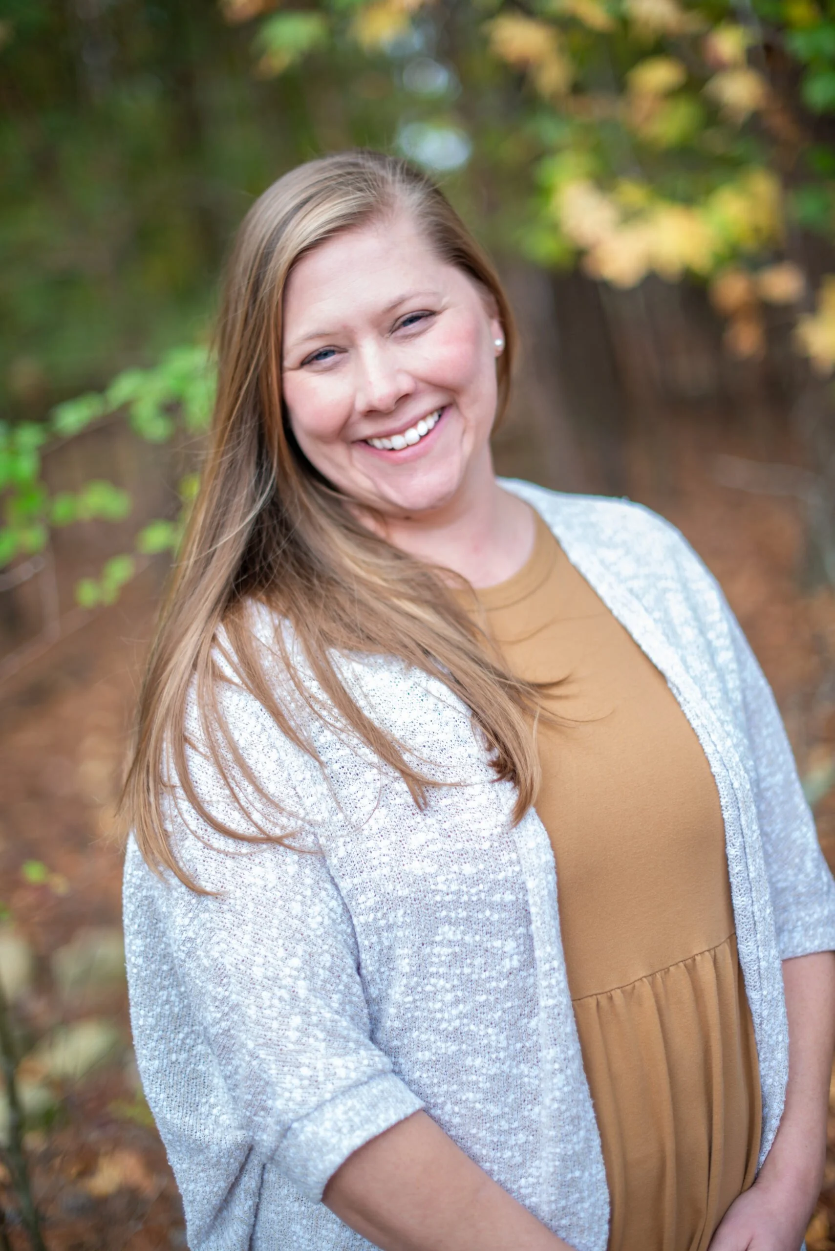 A woman with long light brown hair smiling outdoors in a wooded area.