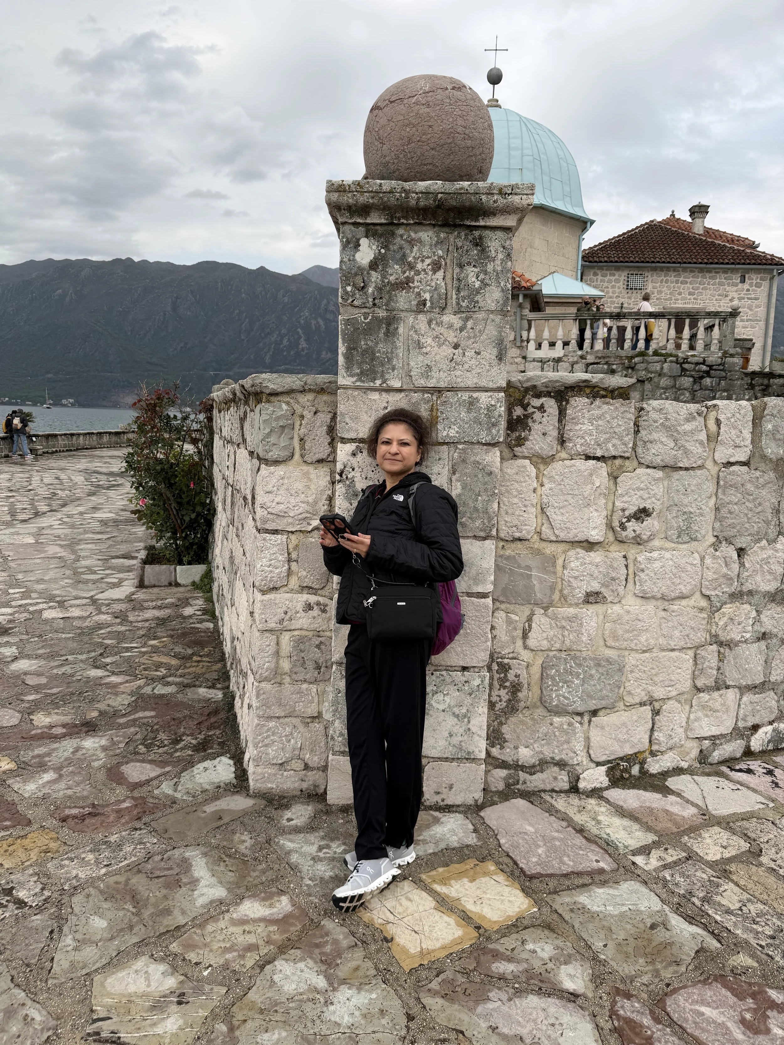 A woman in black pants, a black jacket, and white sneakers stands against a stone wall on a cobblestone path near water and mountains. She holds a phone and has a purple backpack. The background includes a stone building with a blue dome and a small group of people on a balcony.