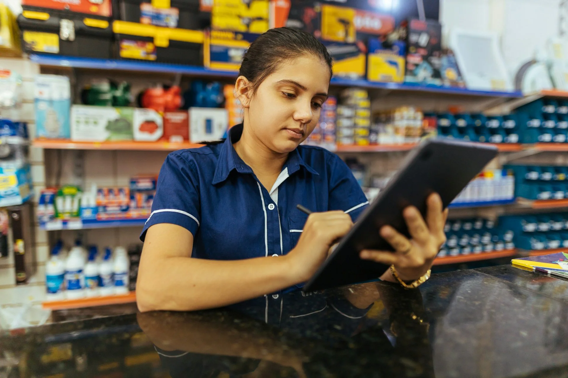 A woman in a blue uniform using a tablet with a stylus at a counter in a hardware store.