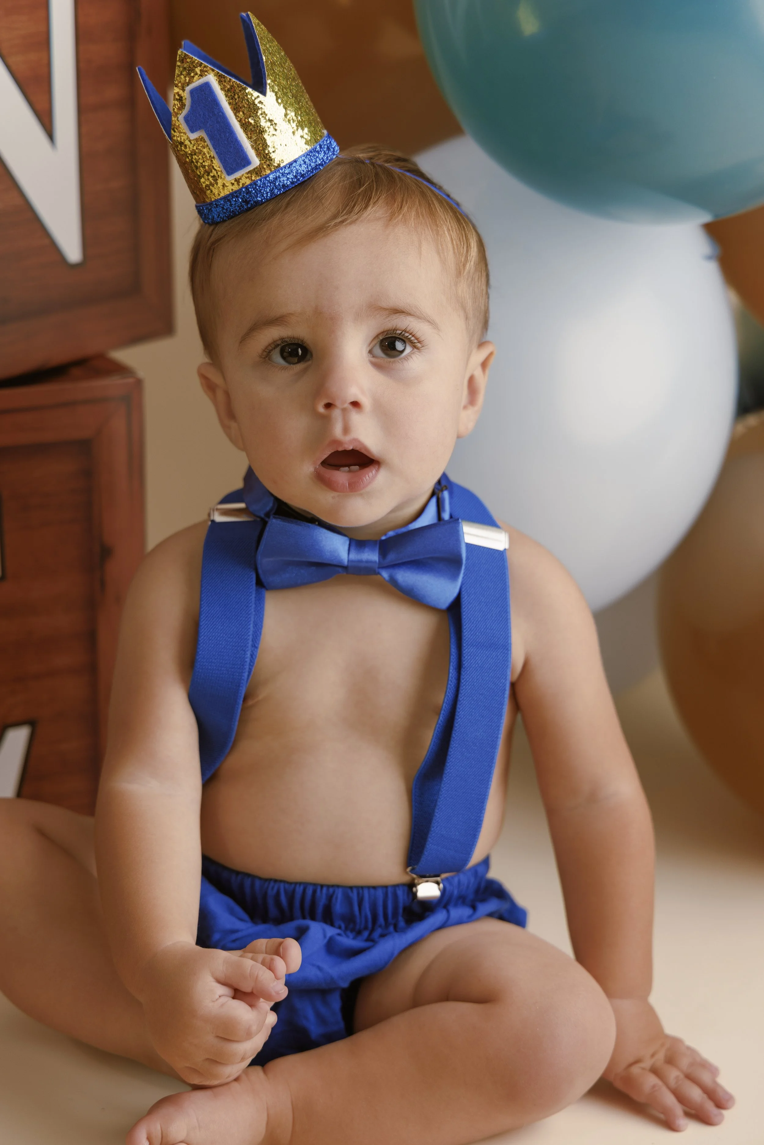 A young child wearing a blue bow tie and suspenders, sitting on an adult's lap, wearing a gold and blue birthday crown with the number 1, and surrounded by balloons.