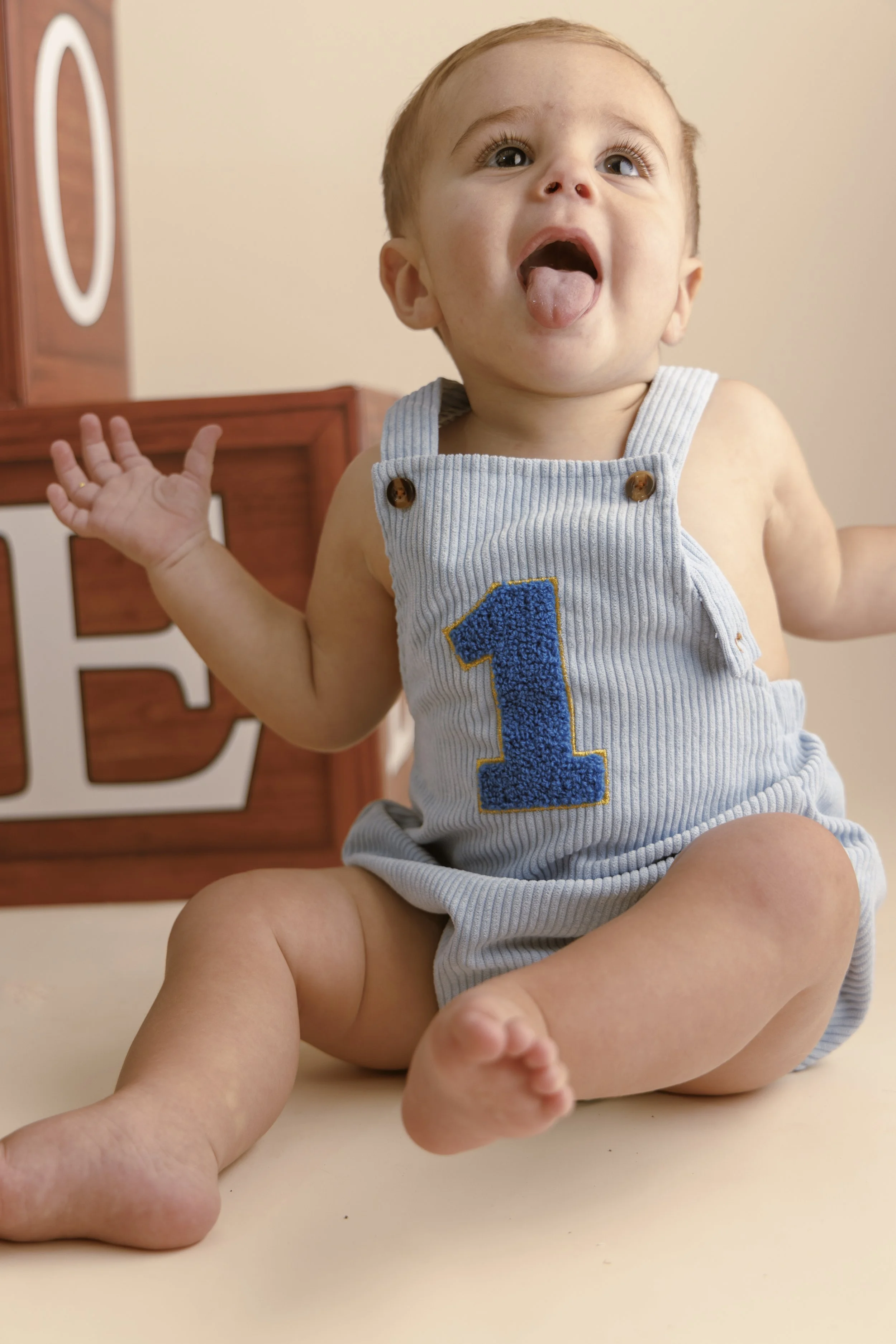 A young child with light skin and short hair sitting on a beige surface. The child is wearing a light blue sleeveless romper with a number one patch on the front. The child's mouth is open, tongue sticking out, and one hand raised. A wooden decoration with large letters is visible in the background.