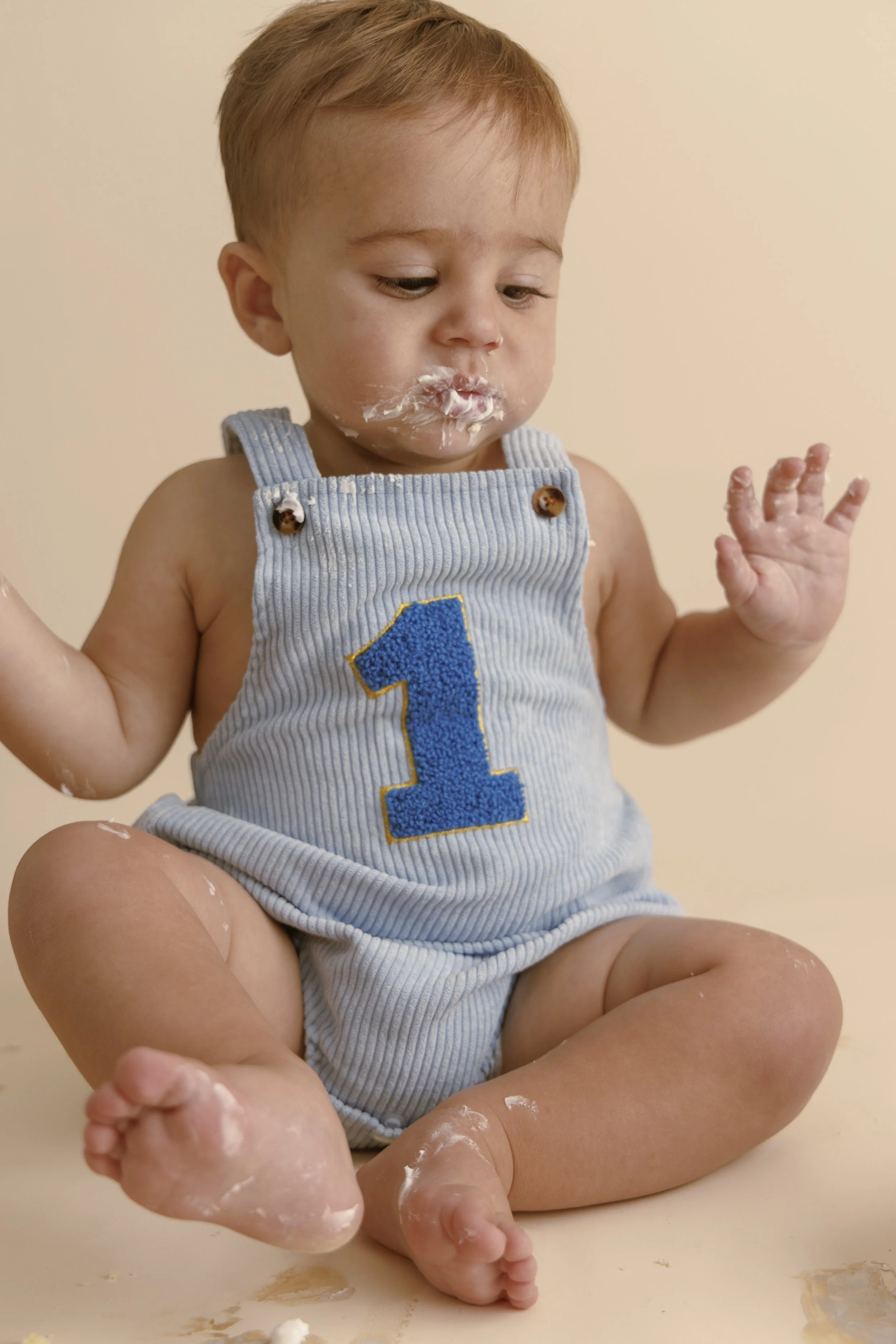 A young child with light skin and red hair sitting cross-legged on a cream-colored surface. The child is wearing a blue and gray sleeveless romper with a large blue number 1 on the front. The child has some white cream or frosting on their face, hands, and legs, and is looking down with a curious expression.