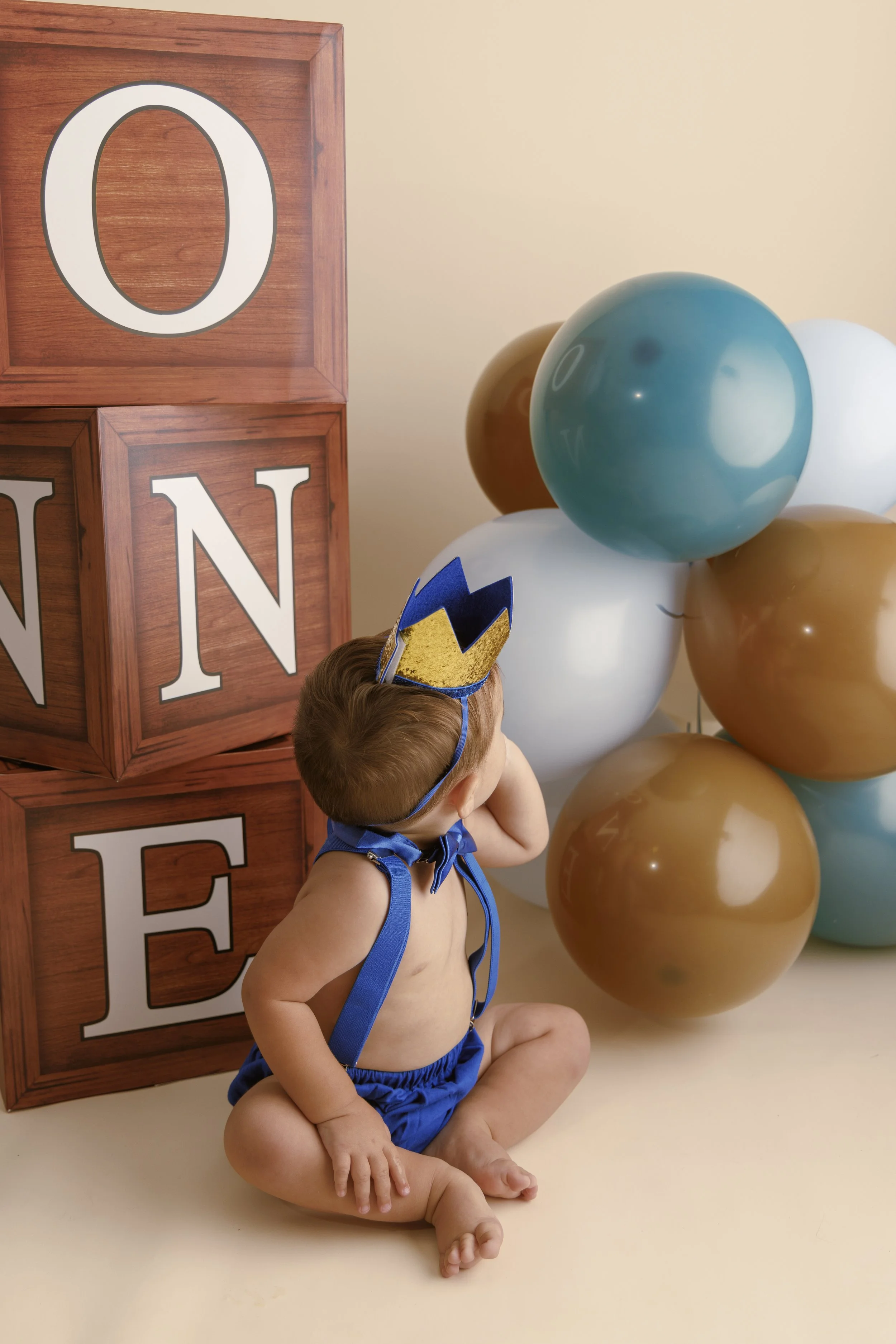 A young child wearing blue shorts with a blue and yellow crown headband, sitting on the floor with arms behind his back, in front of a wooden block decoration and balloons in gold, white, blue, and black. The child is shirtless and appears to be at a celebration or party.