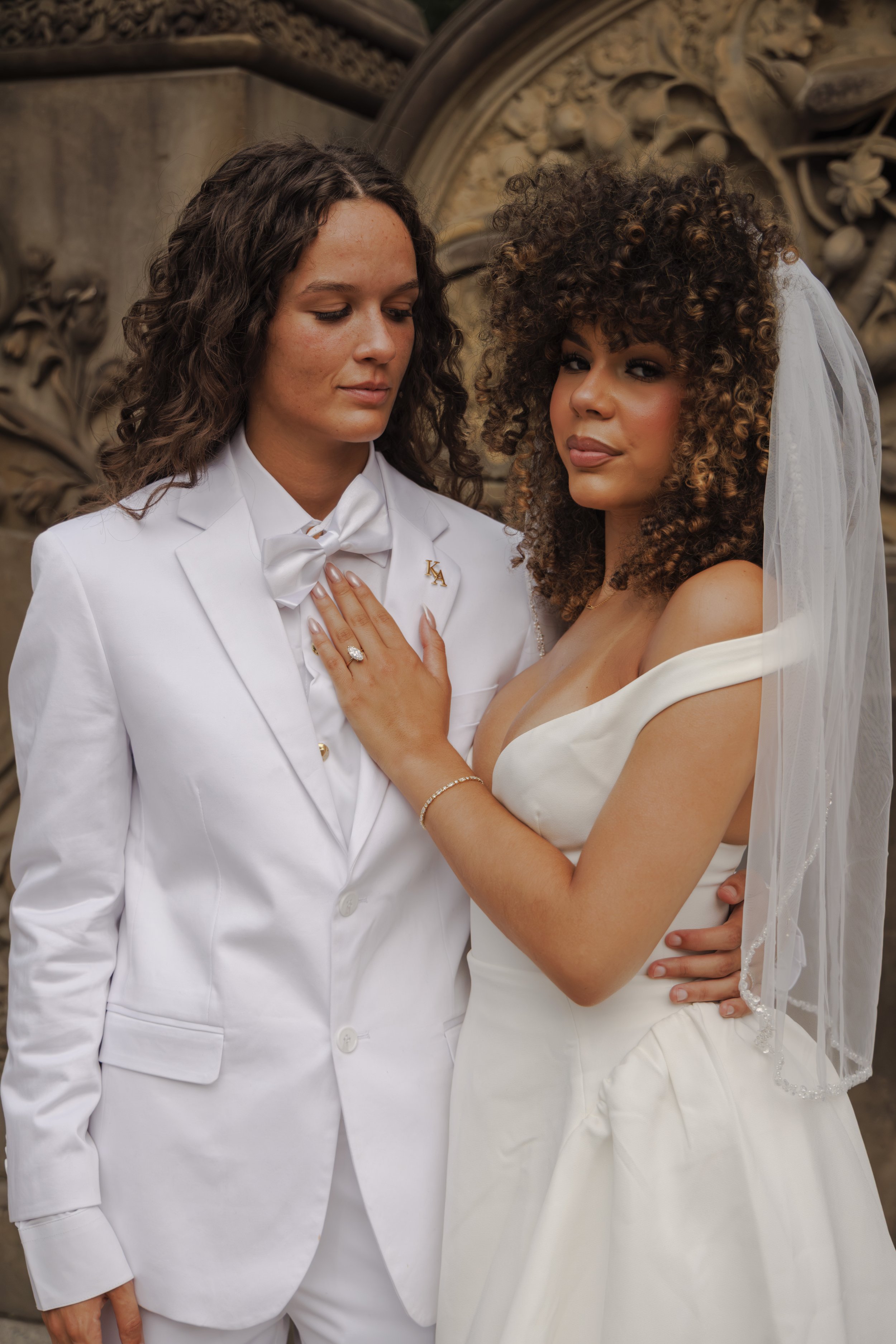 Couple dressed as a bride and groom, standing close together in front of a decorative stone arch, with the bride wearing a white off-shoulder wedding gown and veil, and the groom in a white tuxedo with a bow tie and a lapel pin.
