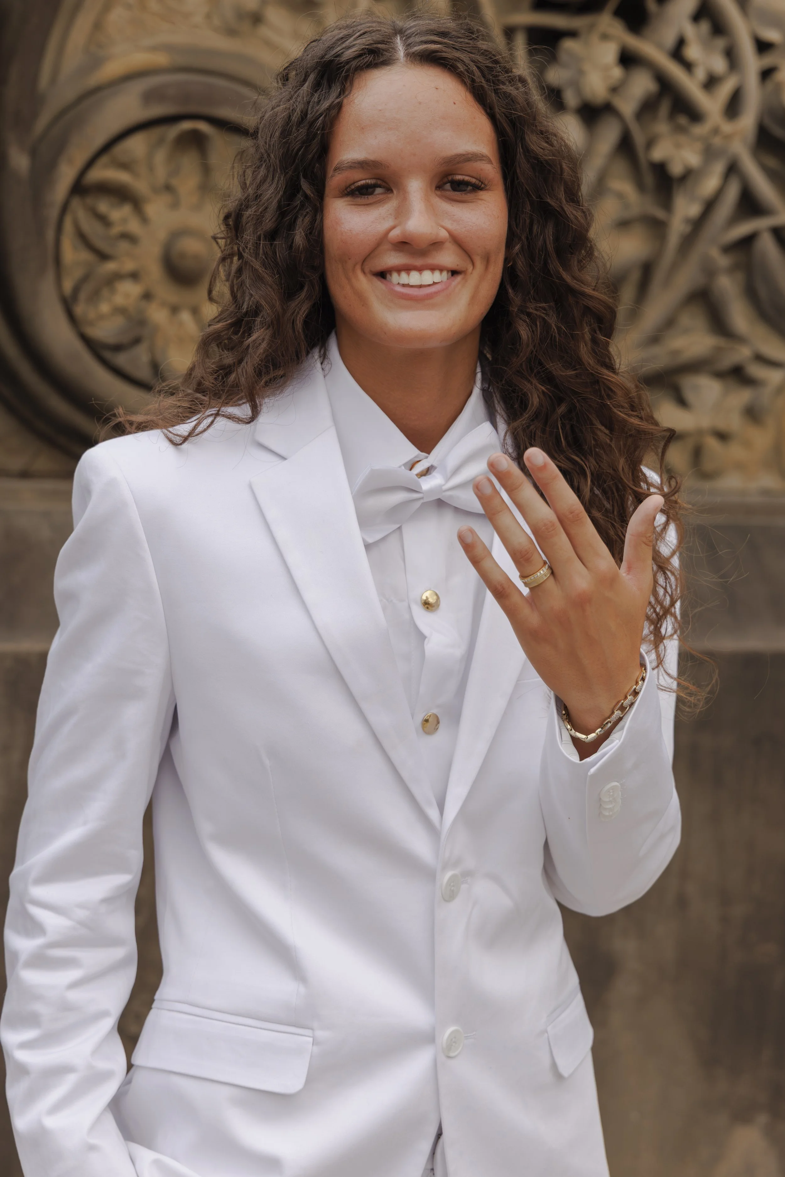 A young woman with long curly brown hair, wearing a white suit with gold buttons and a matching bow tie, smiling and showing a wedding ring on her finger, standing in front of an ornate stone architectural background.