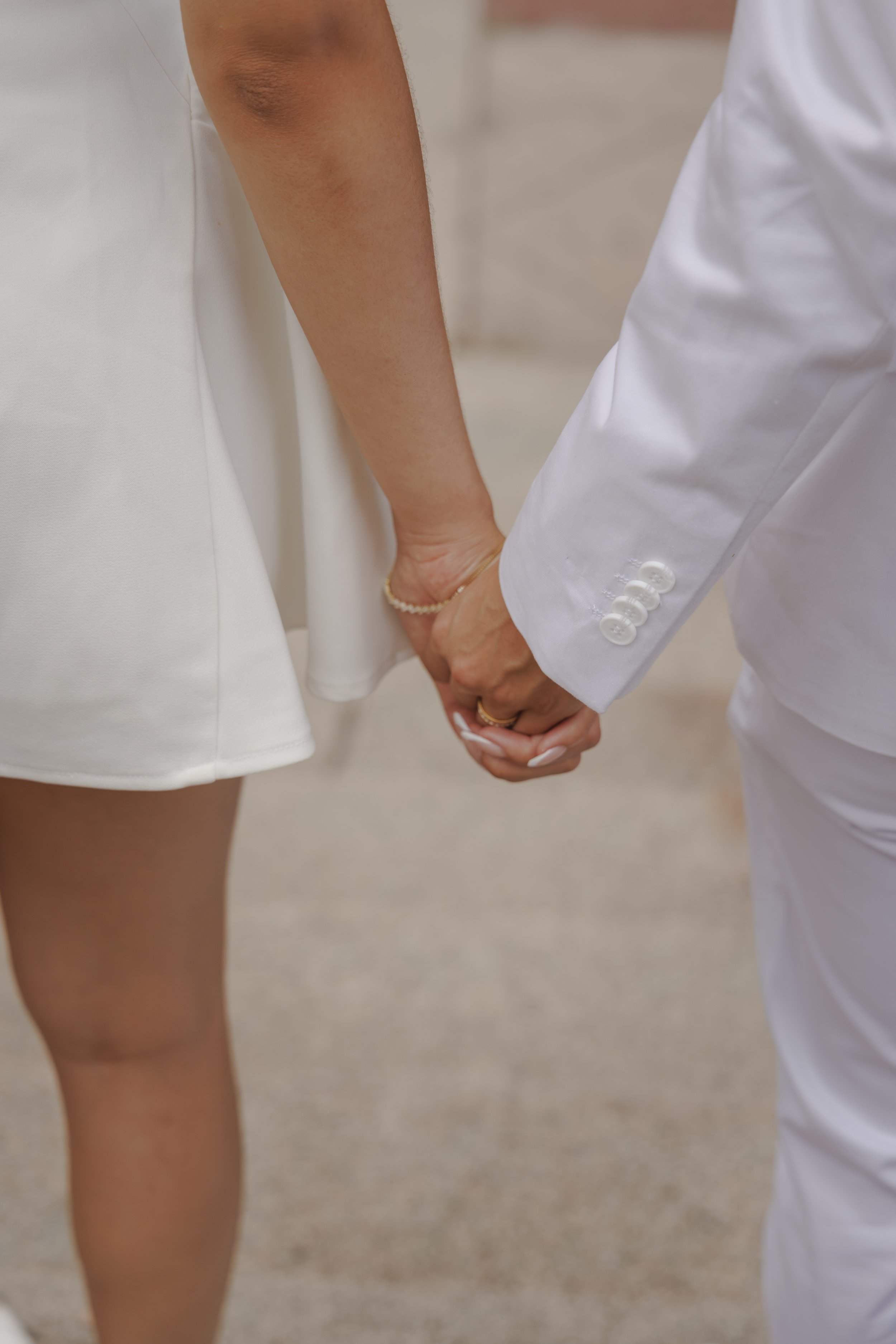A couple holding hands, with the woman's hand on top, wearing a pearl bracelet, and the man dressed in a white suit with button details on the sleeve.