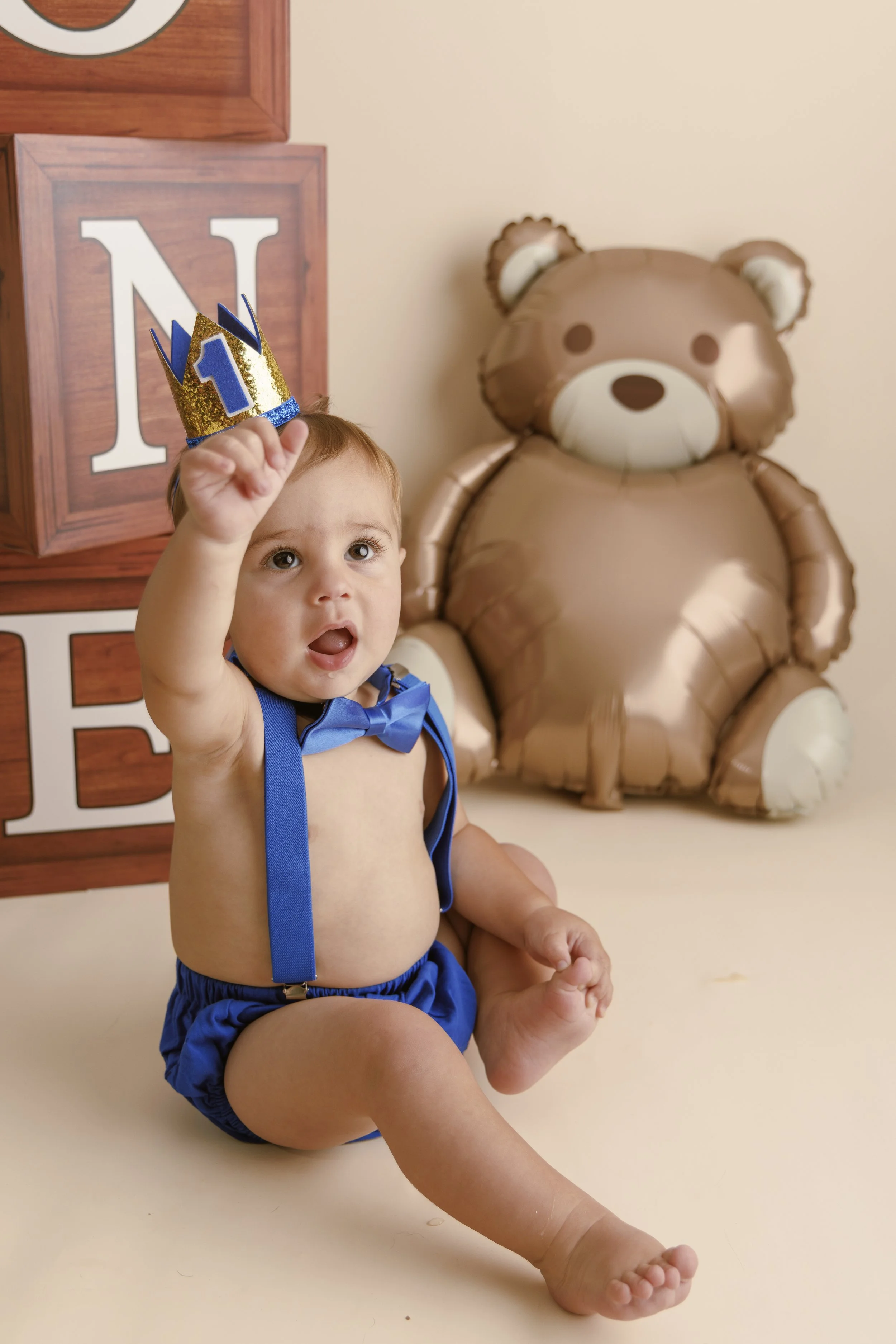A young boy sitting on the floor wearing a blue outfit with suspenders and a bow tie, holding a gold and blue birthday crown with the number 1 on it, with a large teddy bear-shaped balloon in the background.
